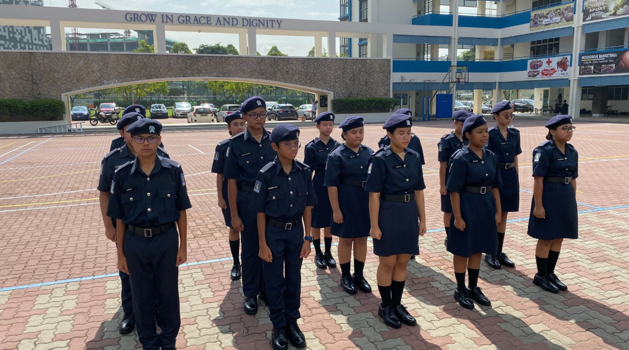 Students in navy uniforms and berets stand in formation on a school courtyard under a "Grow in Grace and Dignity" arch.