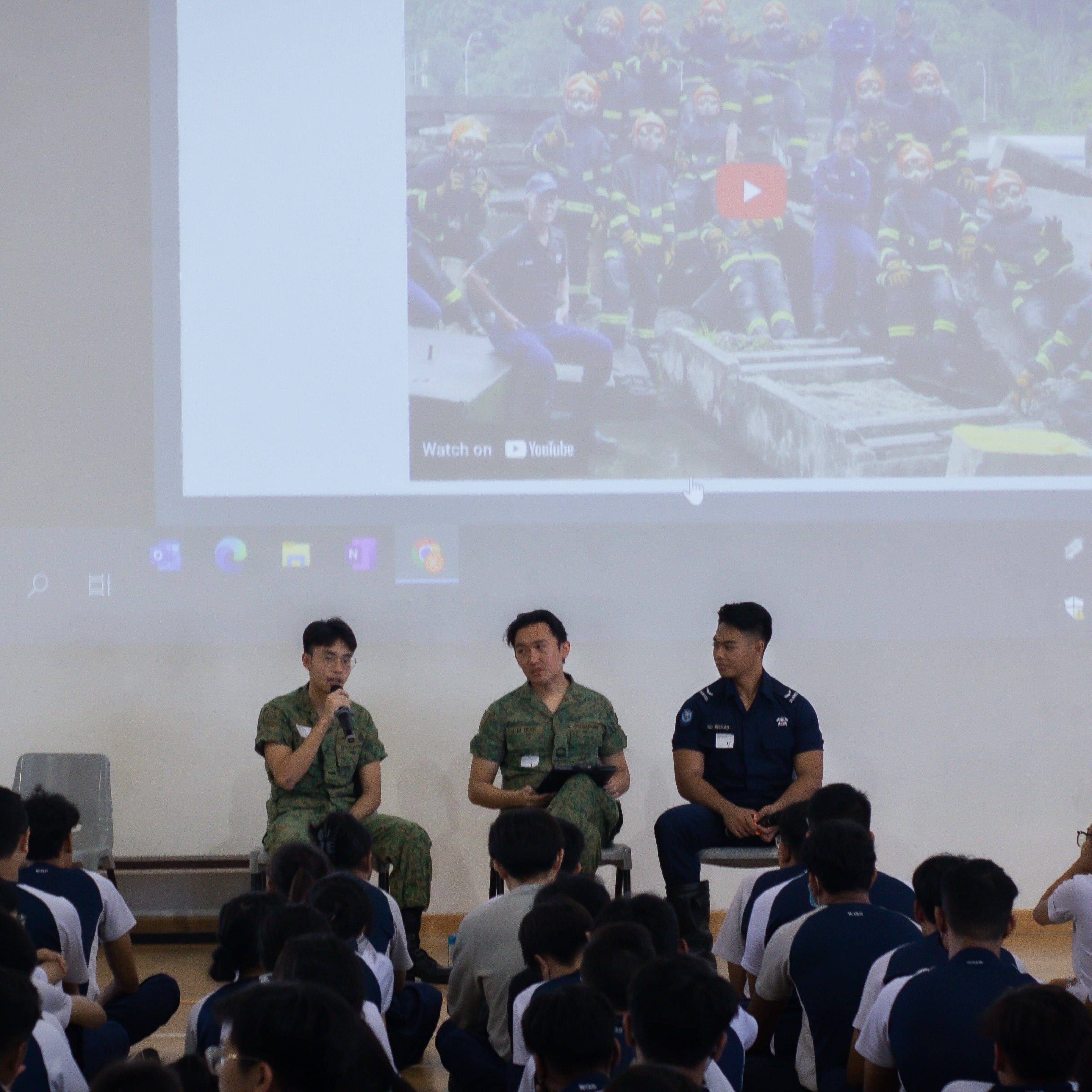 Three uniformed military personnel sit on chairs speaking to a seated audience in front of a YouTube projection.