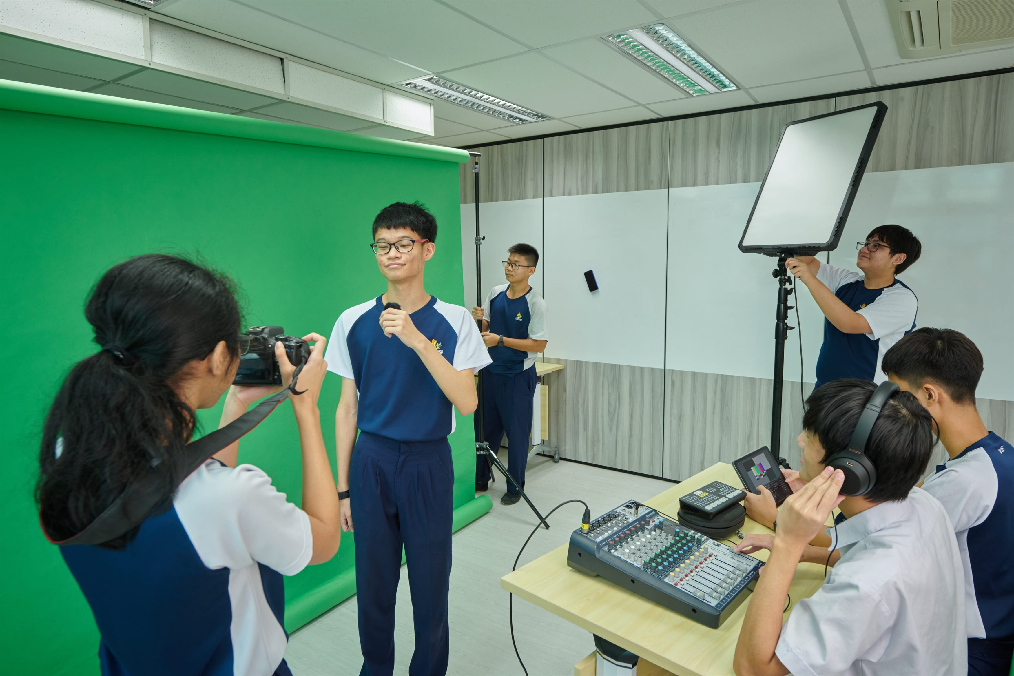 Students in school uniforms film a video in a studio with a green screen, camera, studio light, and audio mixer.