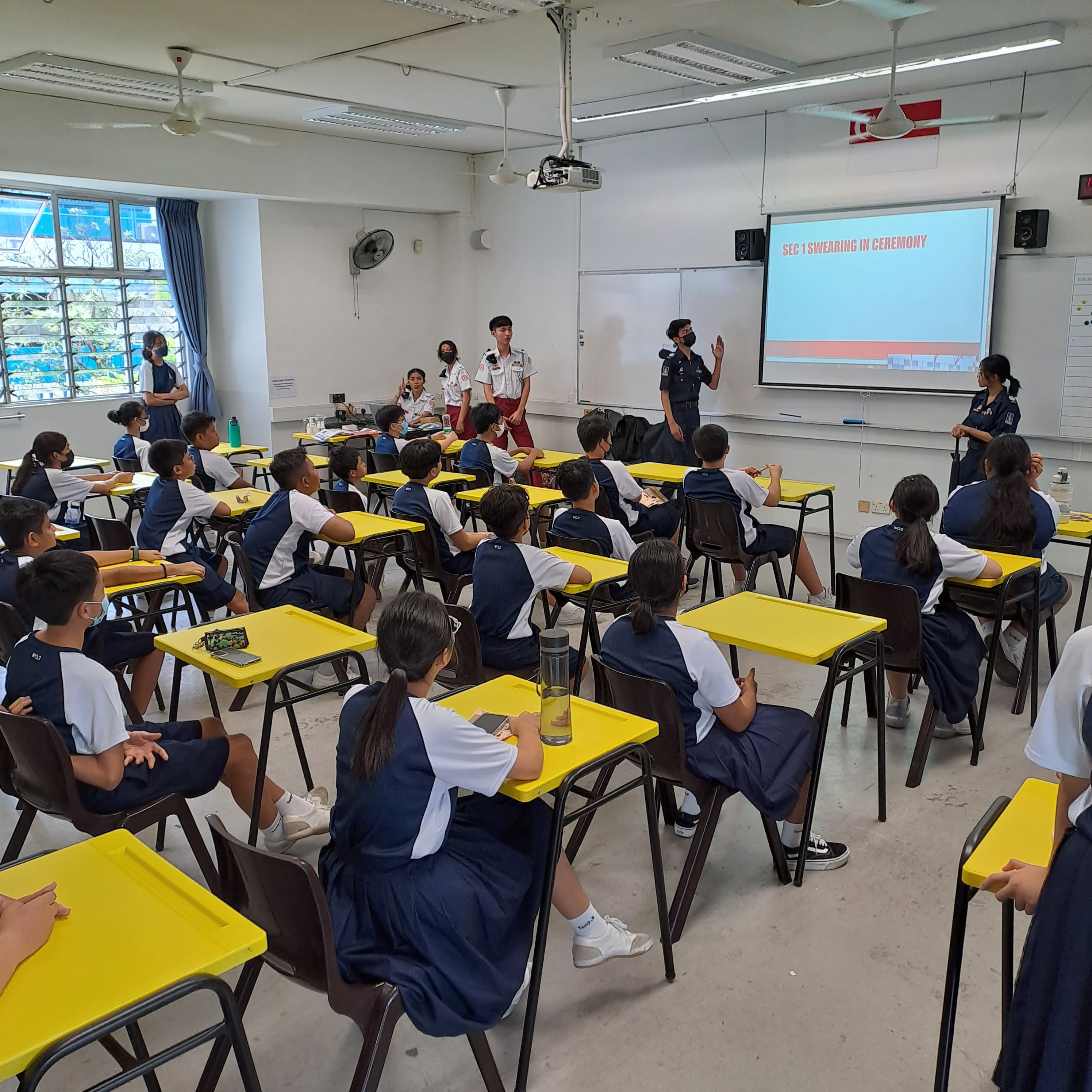 Students in uniform sit at yellow desks facing a screen displaying "SEC 1 SWEARING IN CEREMONY" in a classroom.
