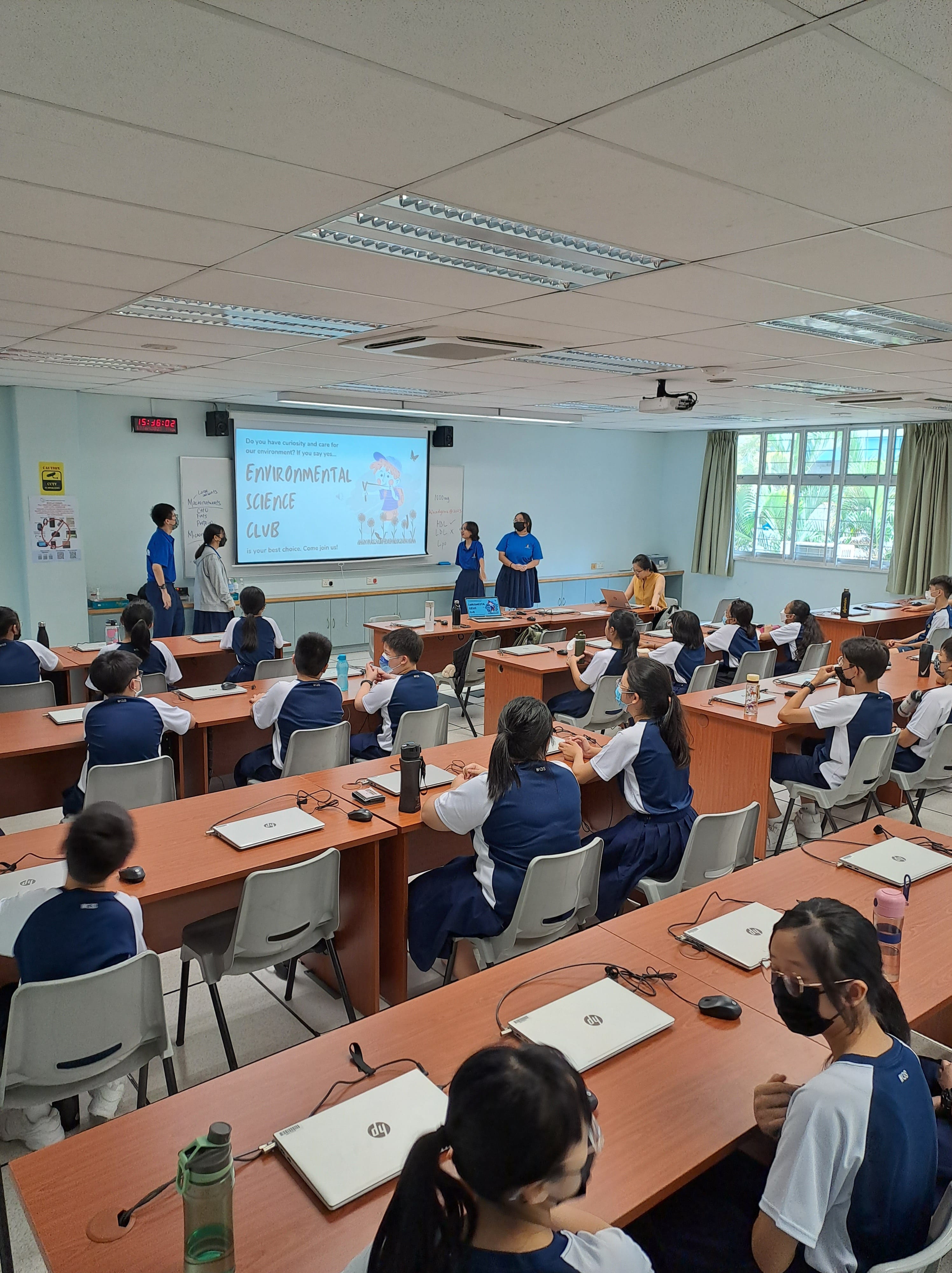 Students in uniform sit at classroom desks watching a projected Environmental Science Club presentation at the front.