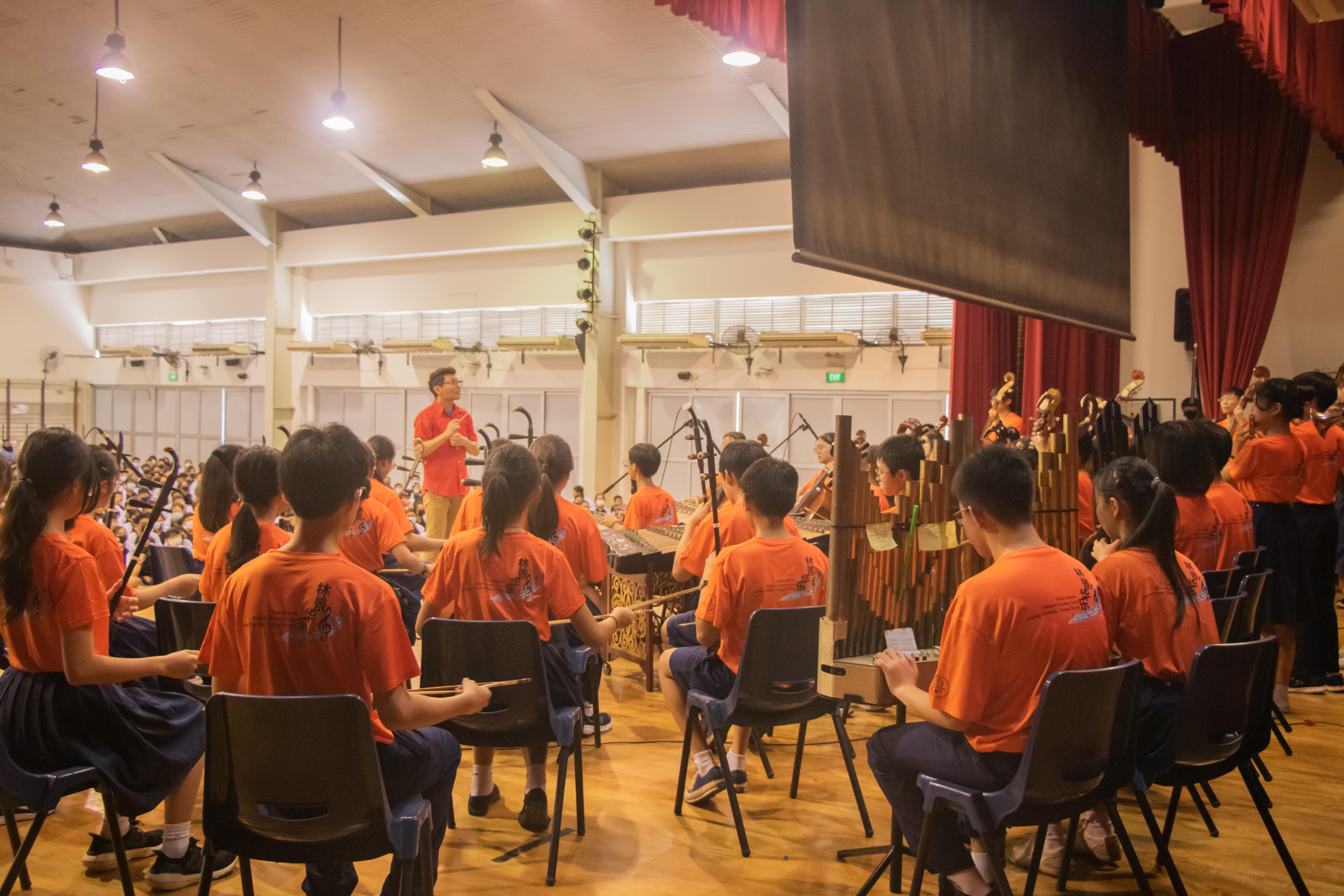 Students in orange shirts play traditional Chinese instruments in a hall, led by a standing conductor in a red shirt.