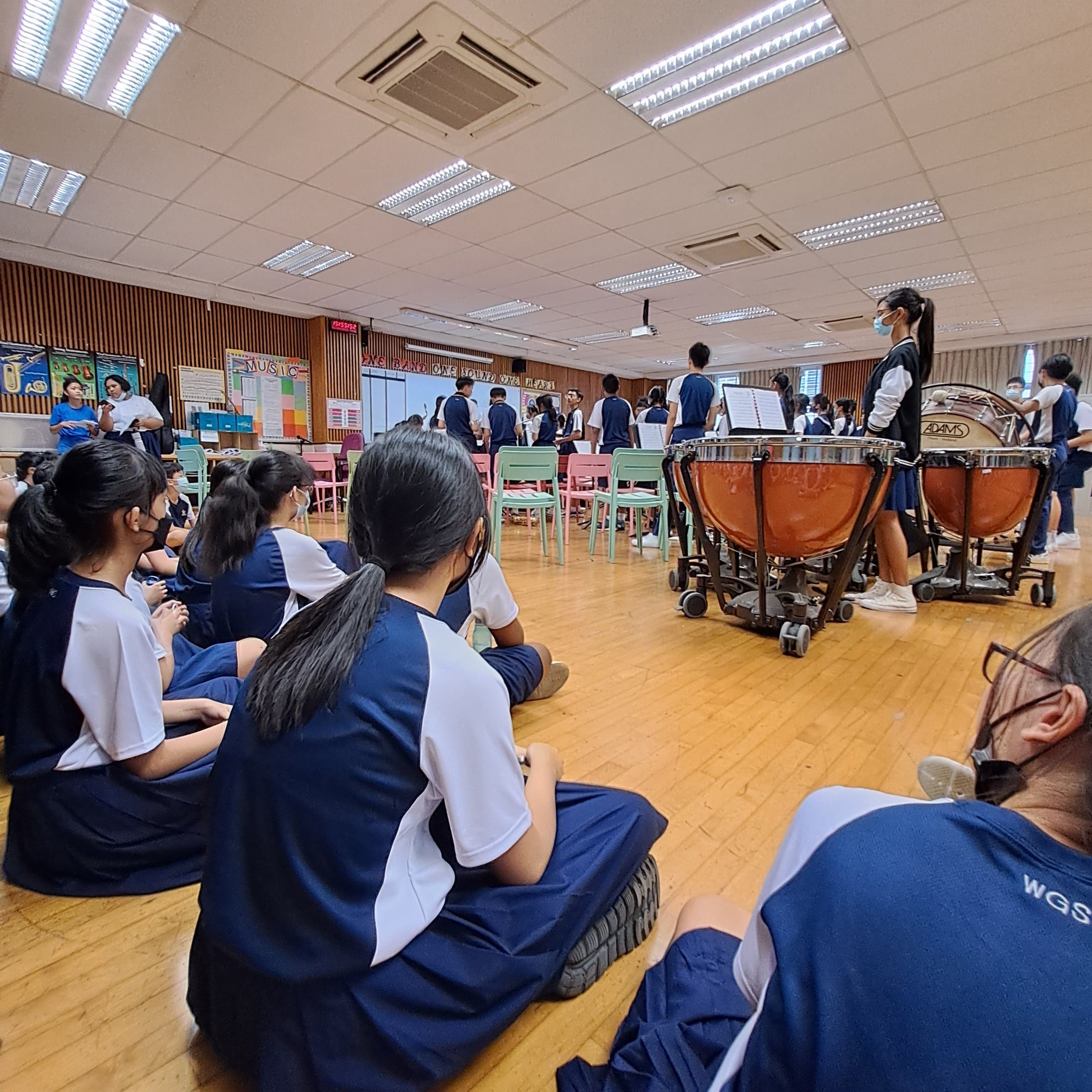 Students in blue and white uniforms sit on a floor watching peers perform near large timpani drums in a school music room.