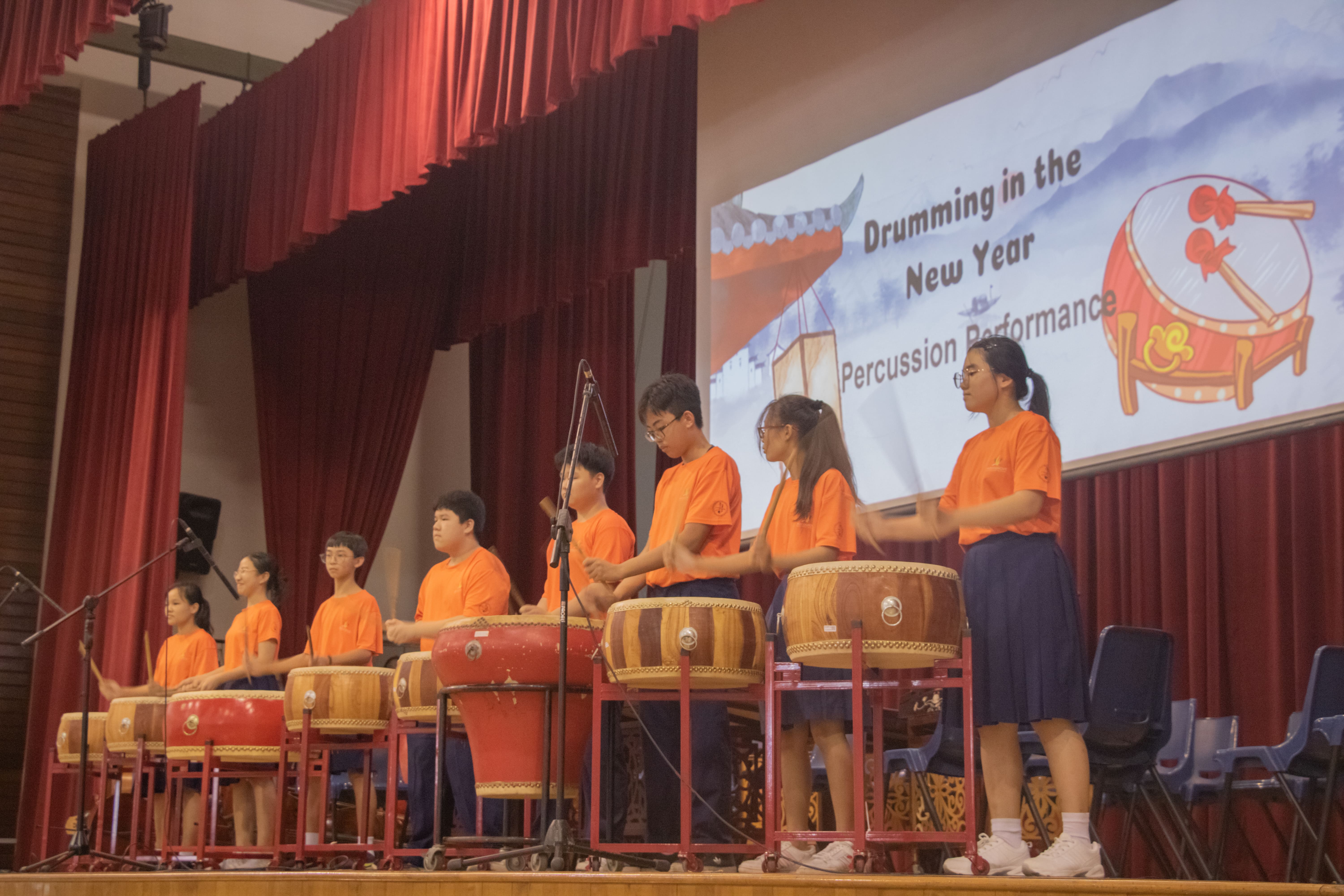 Students in orange shirts perform a New Year percussion show on stage with Chinese drums and a slide reading "Drumming in the New Year."