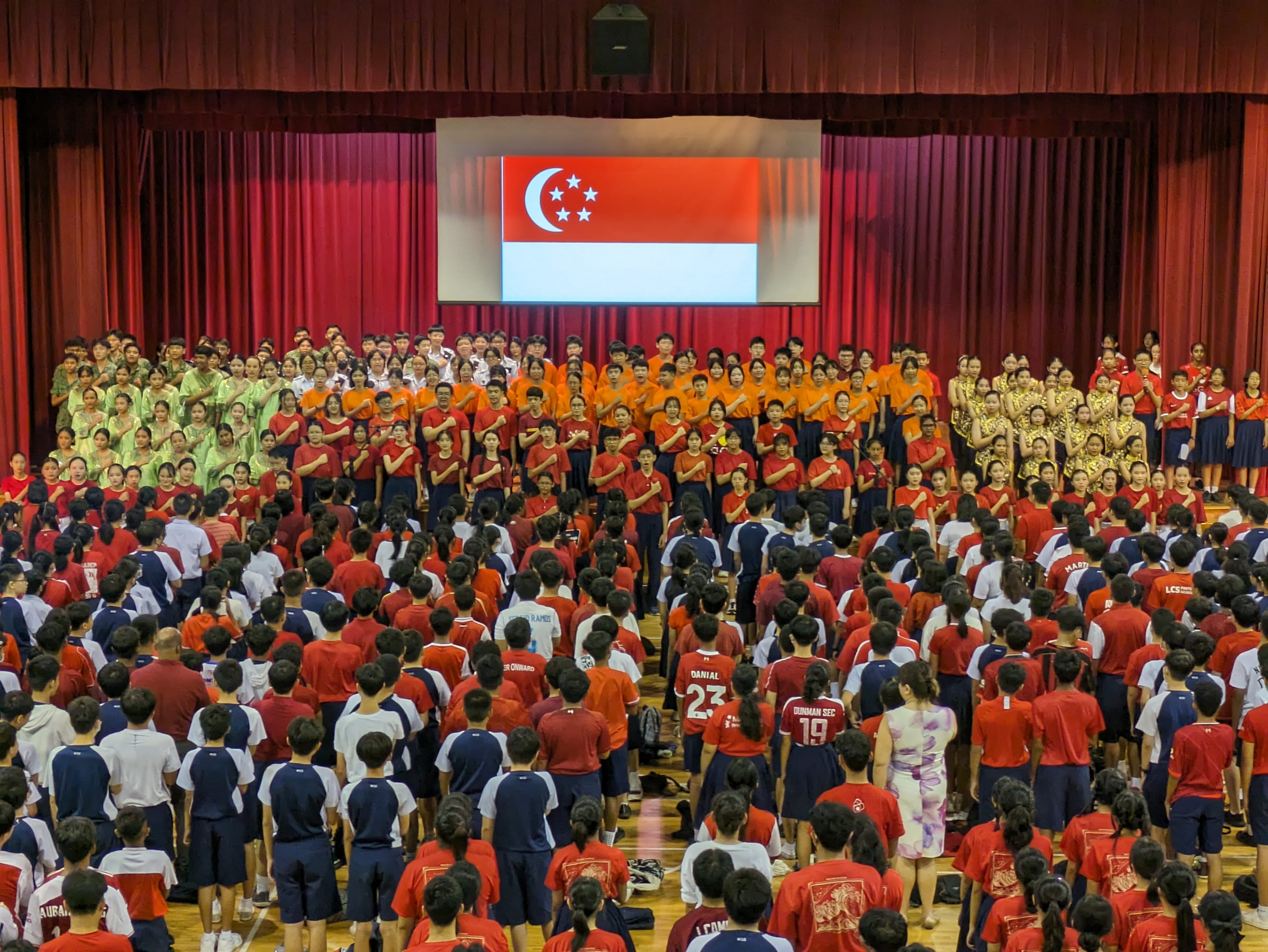 Large crowd of students in a school hall pledging with hands on chest, Singapore flag displayed on stage screen.