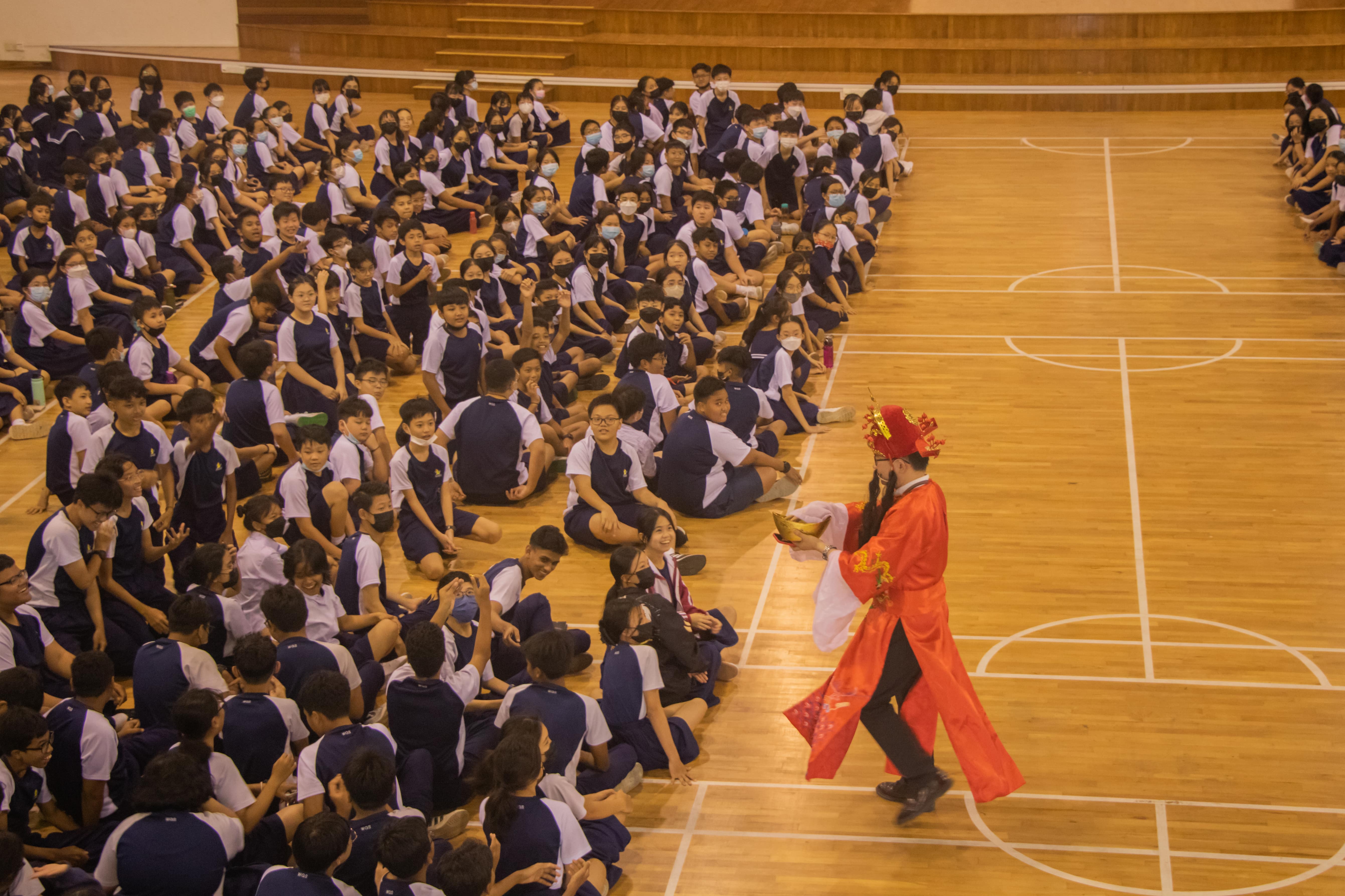 A performer in red traditional Chinese costume walks before students in uniforms seated on a gymnasium floor.