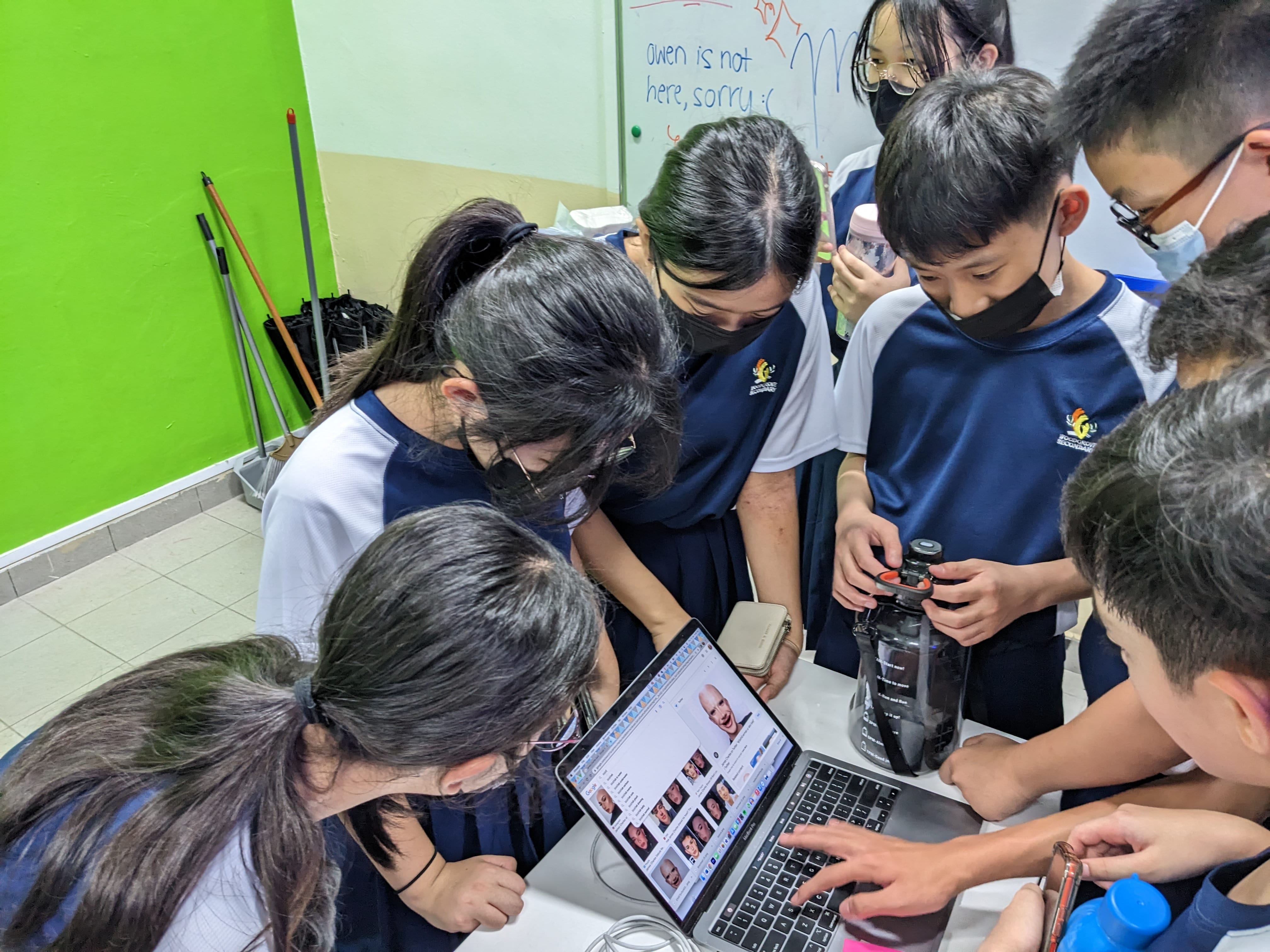 Several school students huddle around a laptop displaying face images in a classroom.