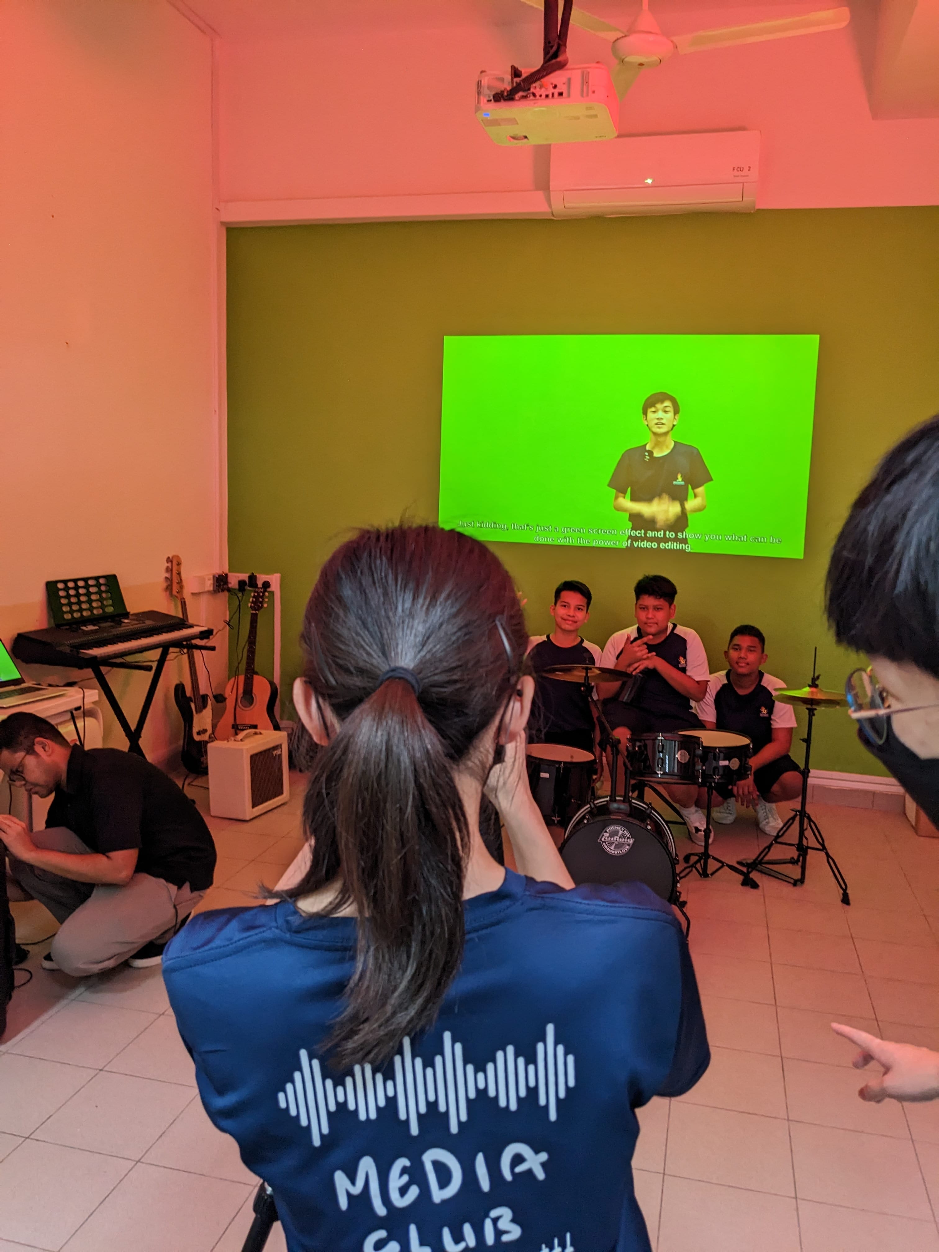 A Media Club member films students at a drum kit while a green screen video plays on a projected screen behind them.