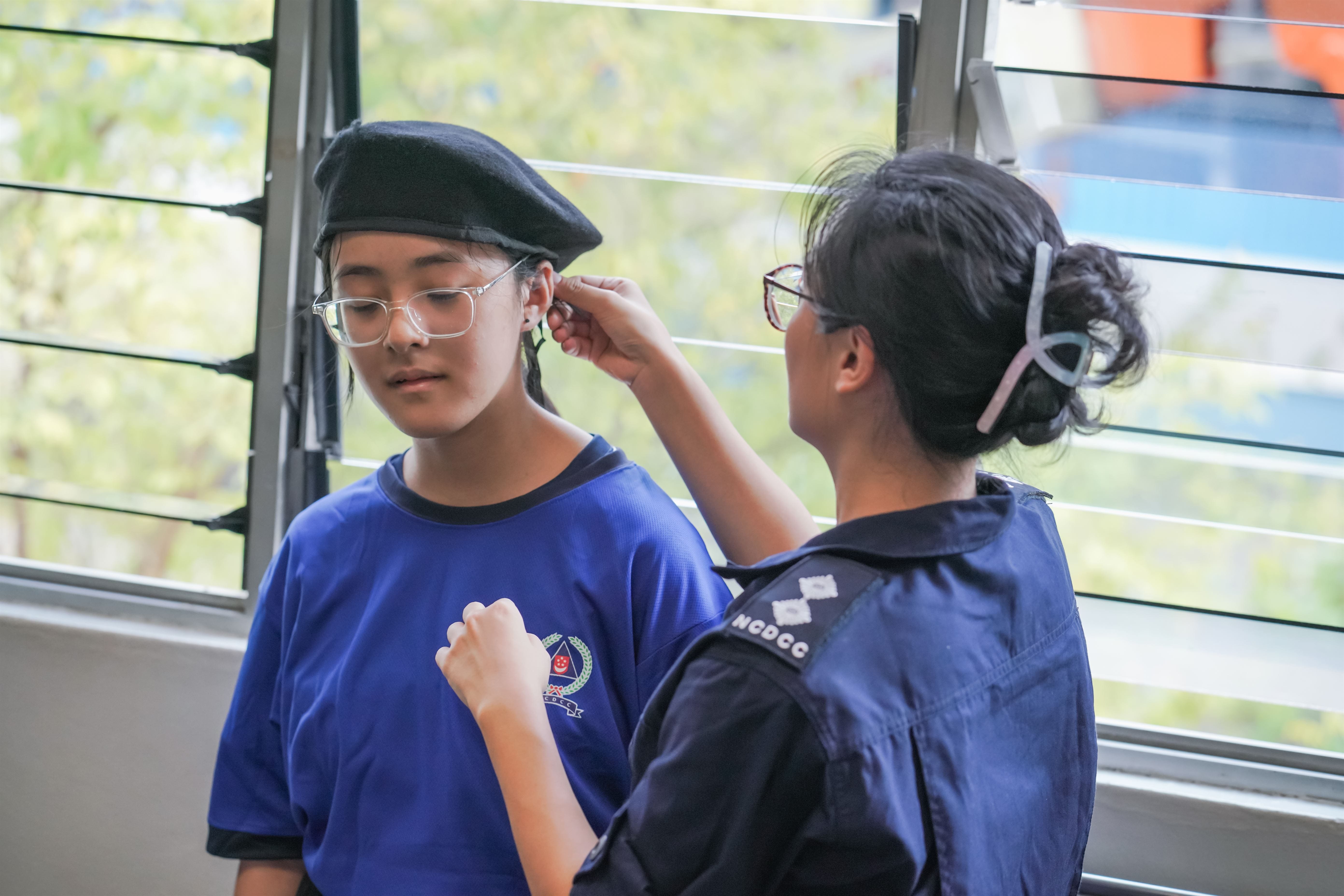 A girl in NCDCC uniform adjusts a black beret on a younger girl wearing glasses and a blue shirt.