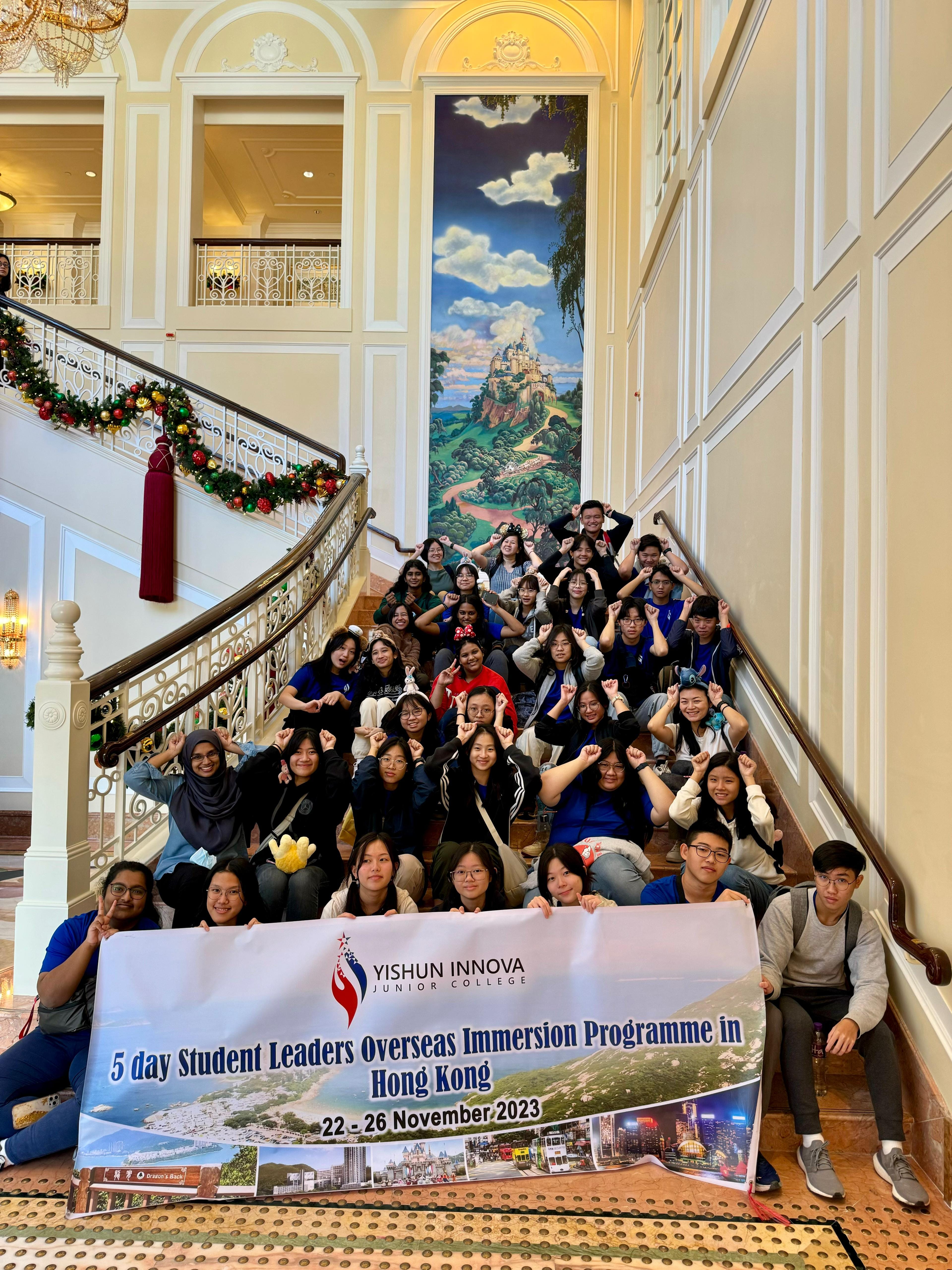Large student group poses on an ornate staircase holding a Yishun Innova Junior College Hong Kong immersion programme banner.