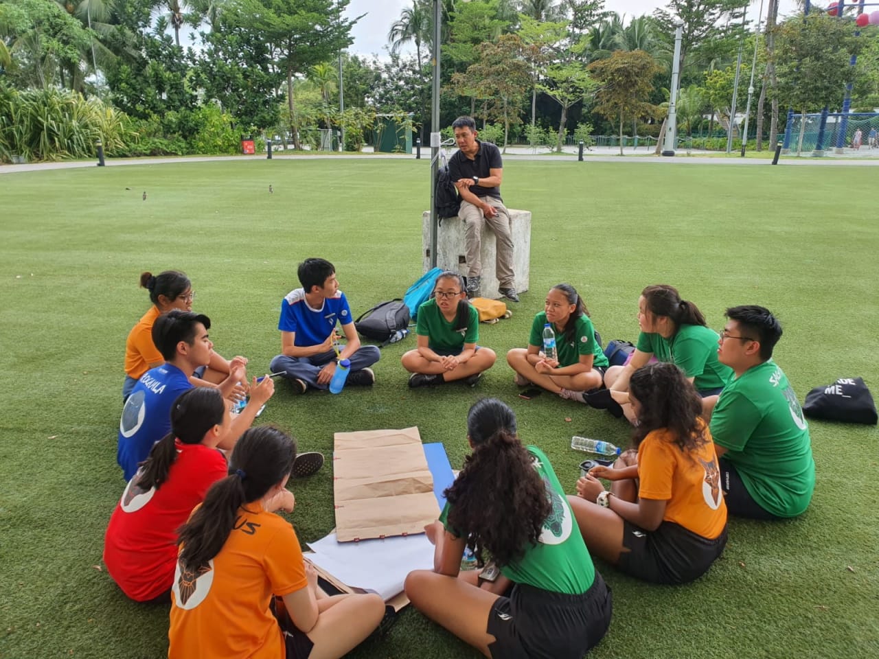 A group of YIJC student leaders sit in a circle on a green field listening to an adult leadership facilitator seated on a block outdoors.