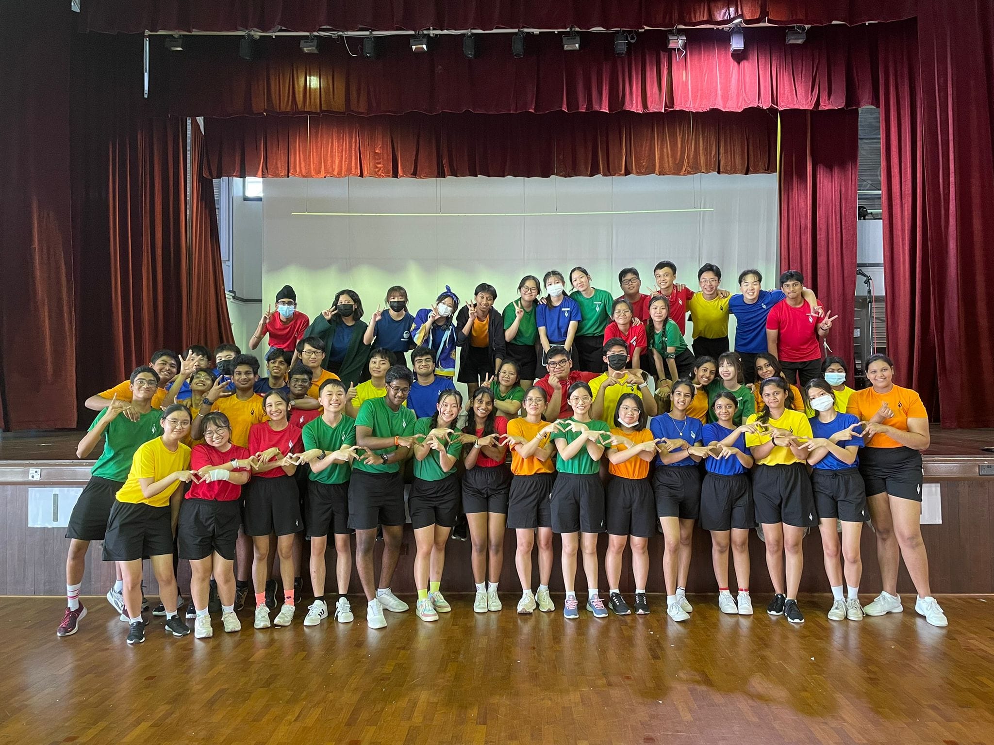Large group of Student Councillors in colorful shirts posing with heart hand gestures in front of a red stage curtain.