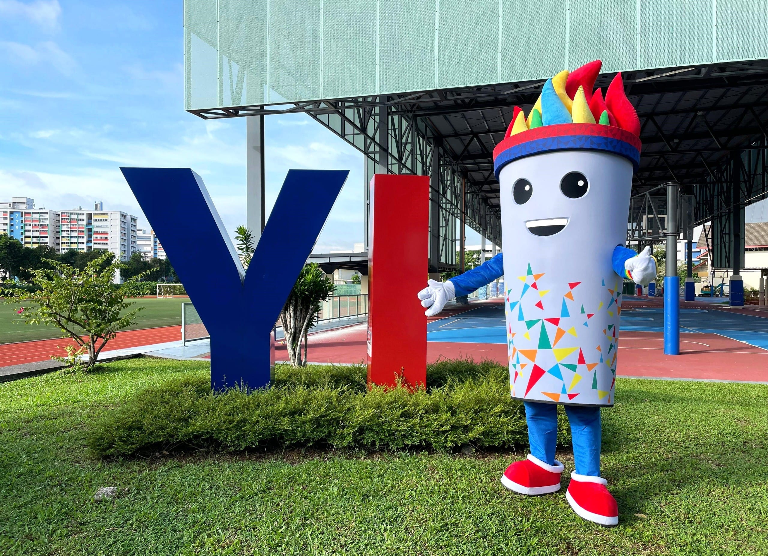 A colorful torch-shaped mascot with a flame head stands beside large blue Y and red I letters at a sports venue.