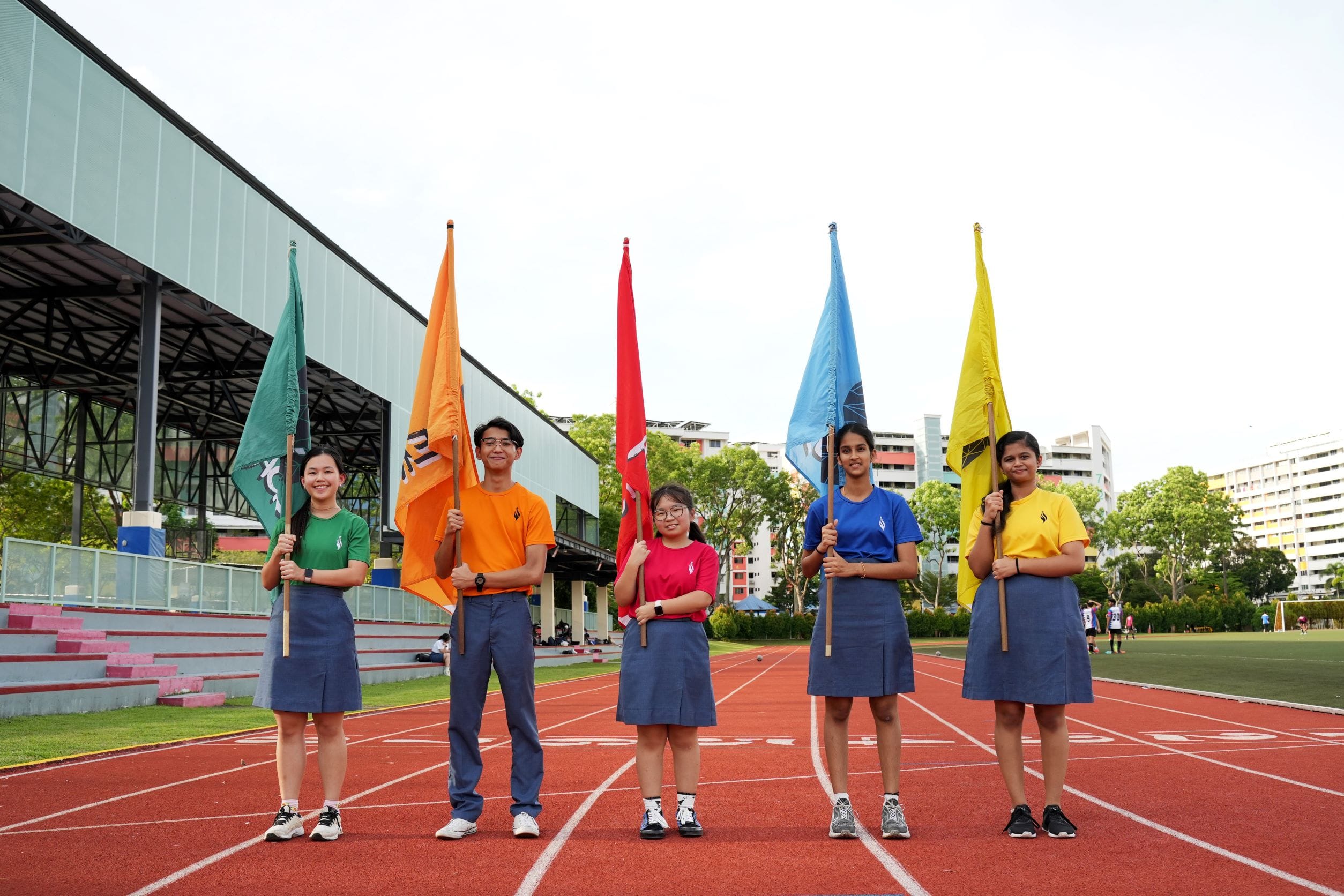 Five students on a running track hold tall flags in green, orange, red, blue, and yellow.