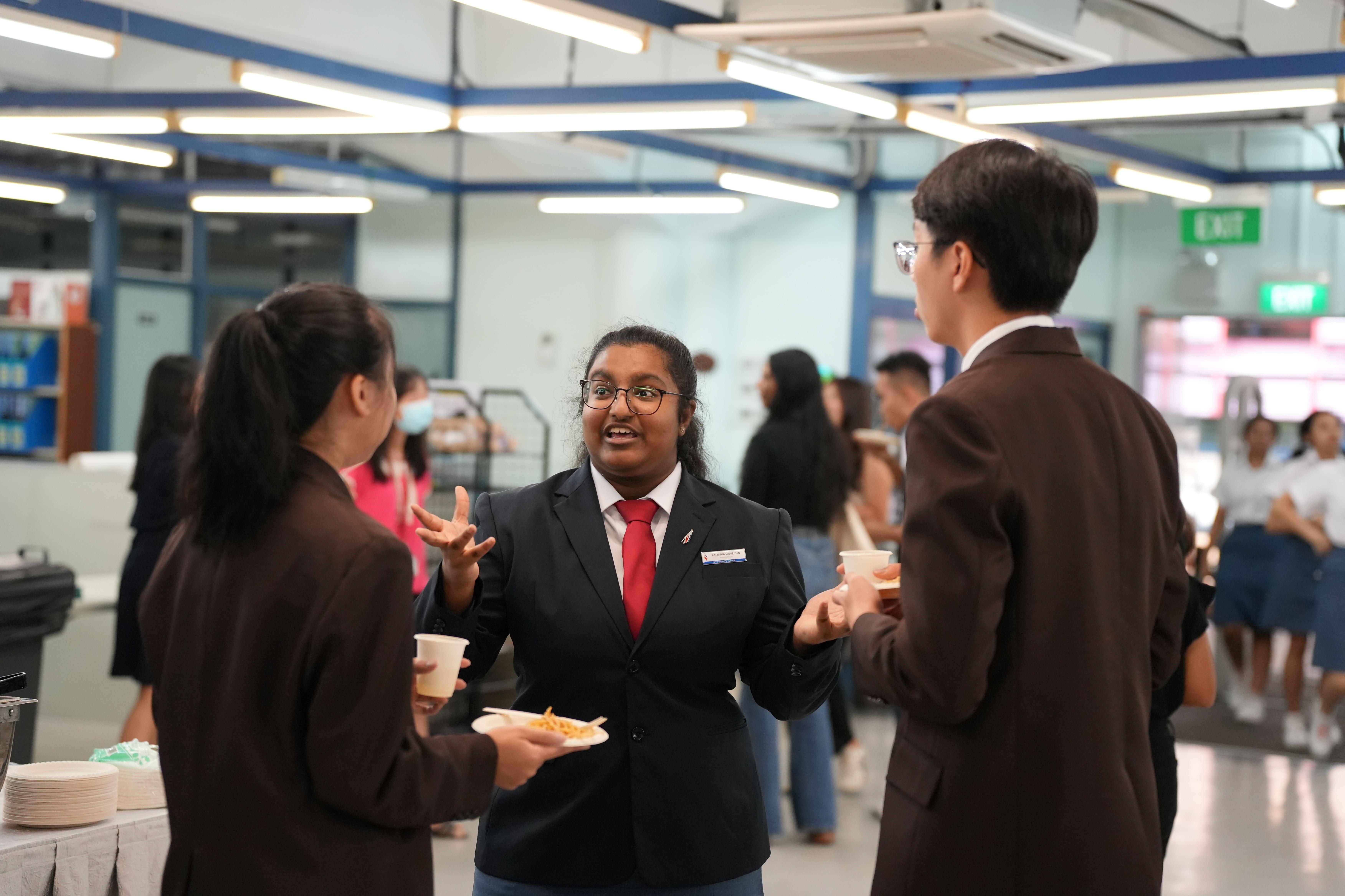 Three student leaders in formal attire chat at an indoor event, holding drinks and a plate of food.