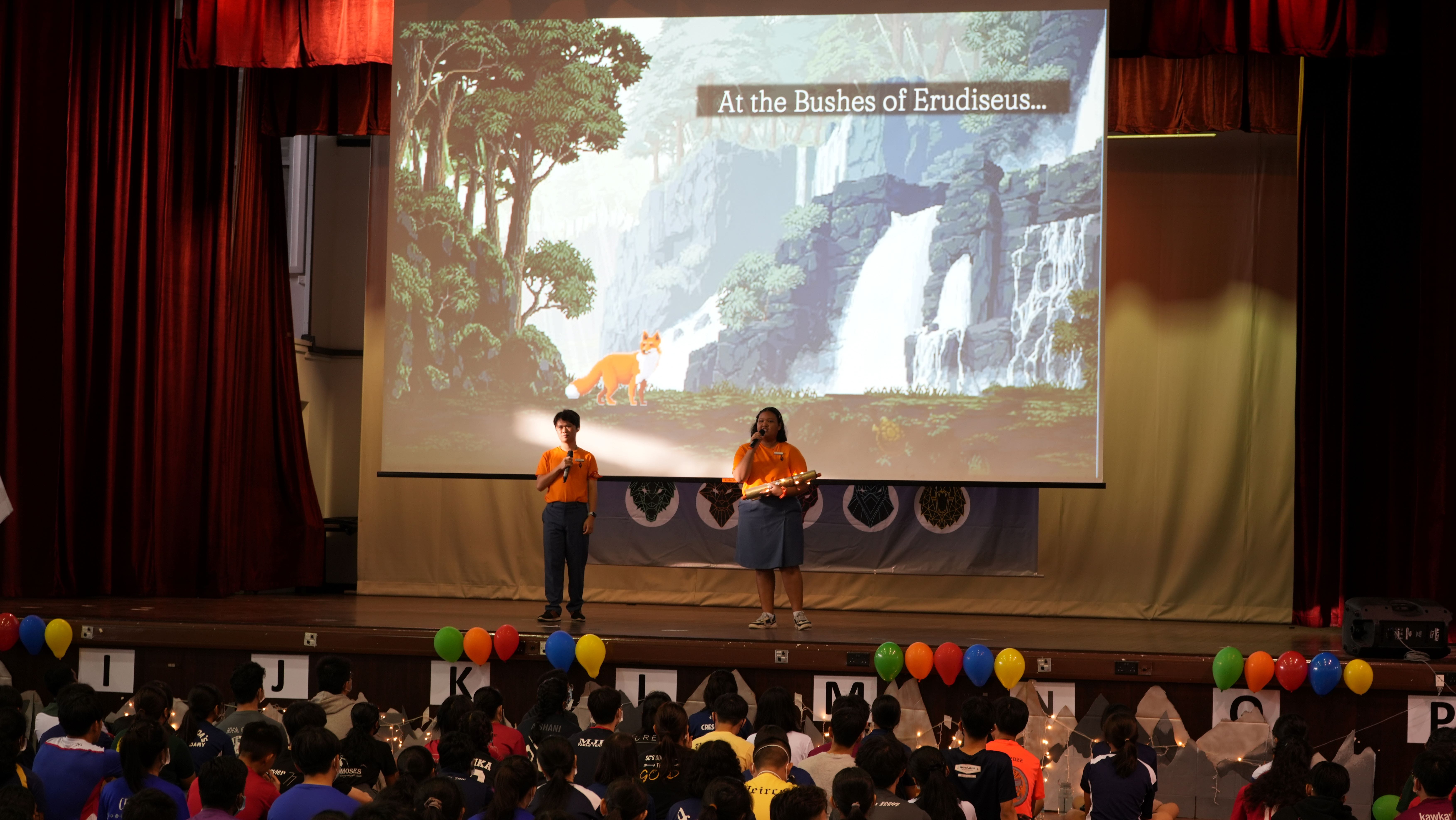 Two presenters in orange shirts on stage with a projected game scene reading "At the Bushes of Erudiseus" before a seated audience.