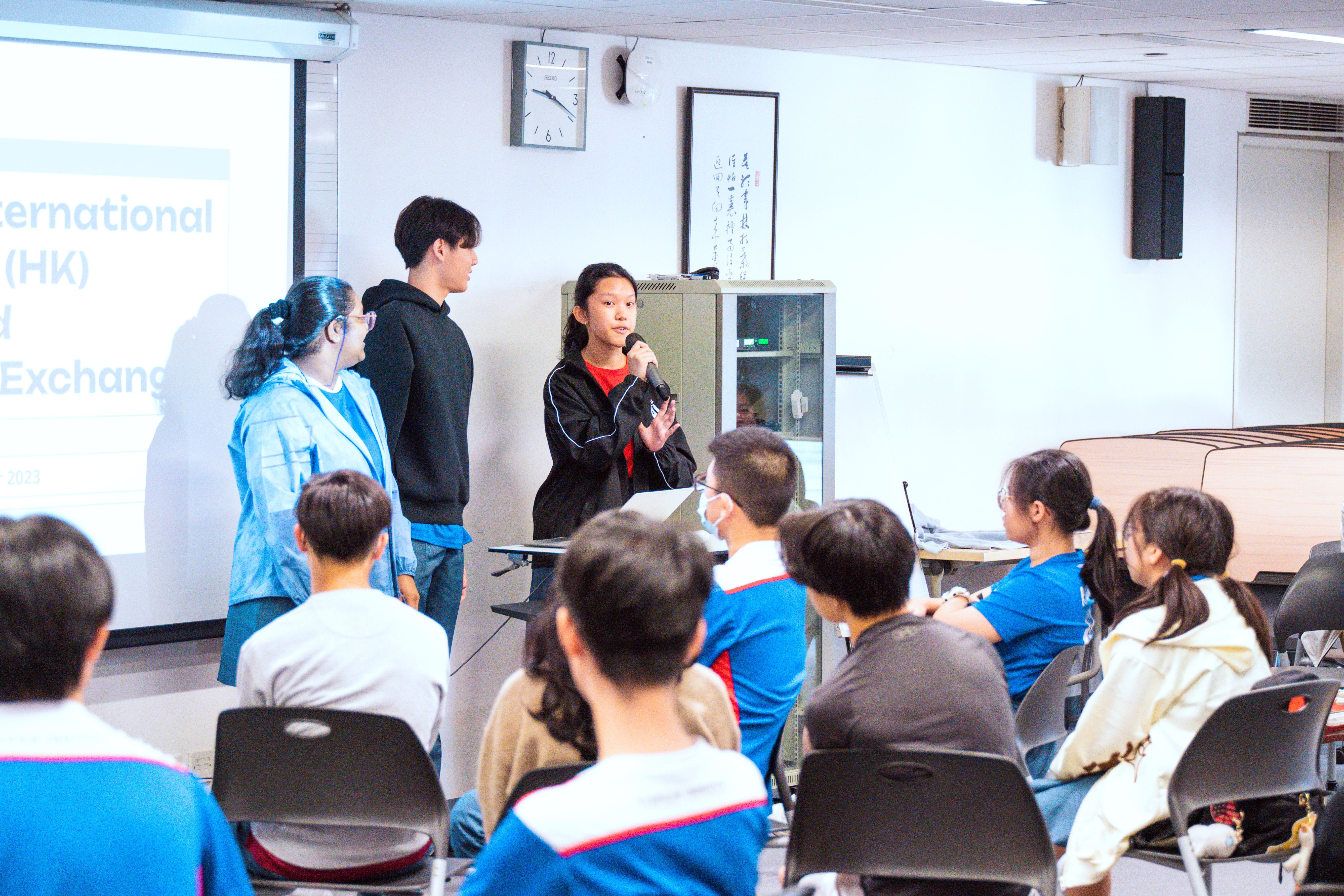 At the overseas Student Leadership Enrichment Programme, a female student leader speaks into a microphone at the front of a classroom while peers sit watching and a slideshow displays behind.