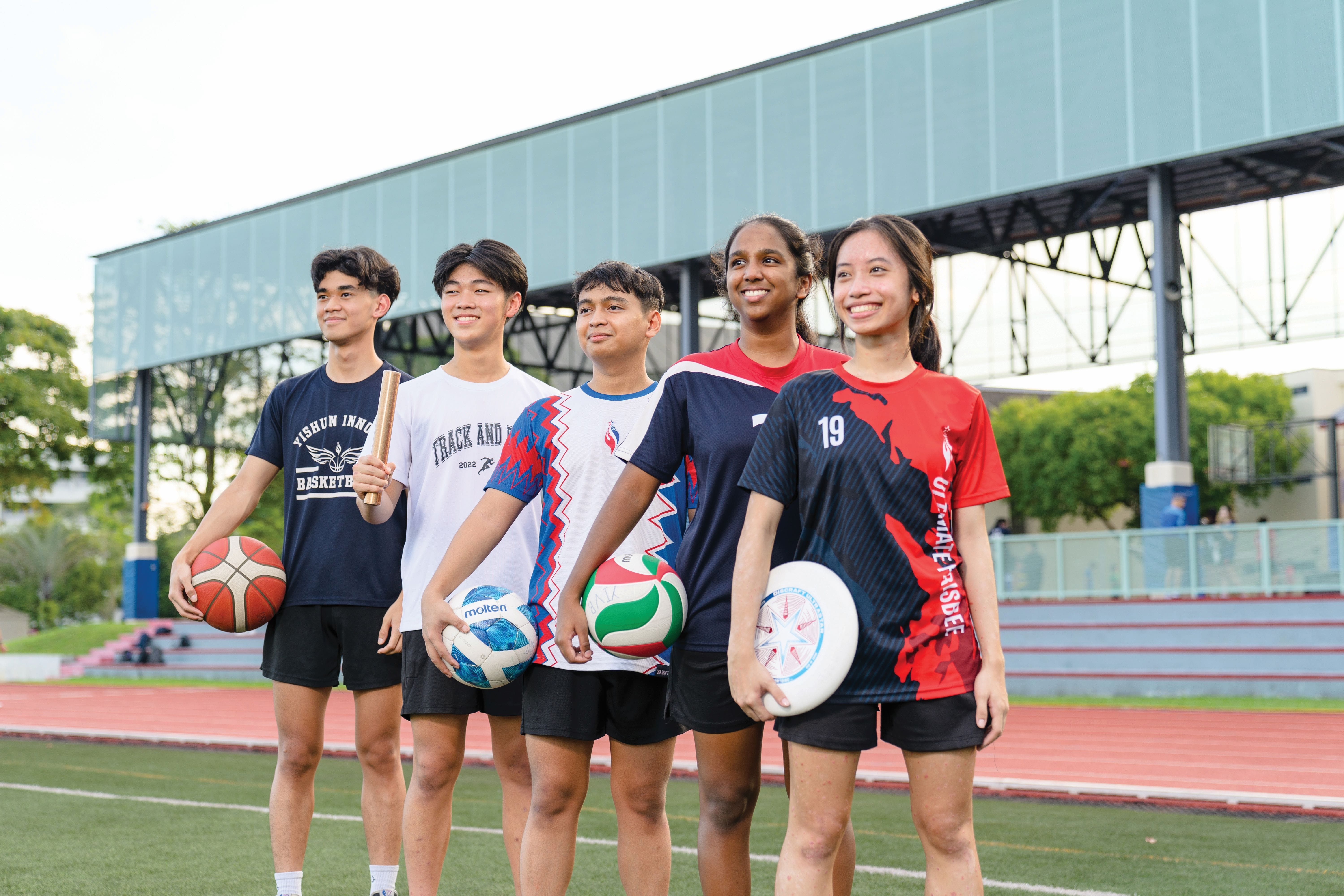 Five smiling student athletes stand on a track field holding a basketball, relay baton, soccer ball, volleyball, and frisbee.