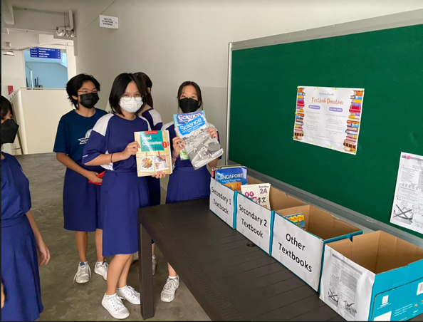 Three masked students in blue uniforms hold textbooks beside labeled donation boxes near a green board.