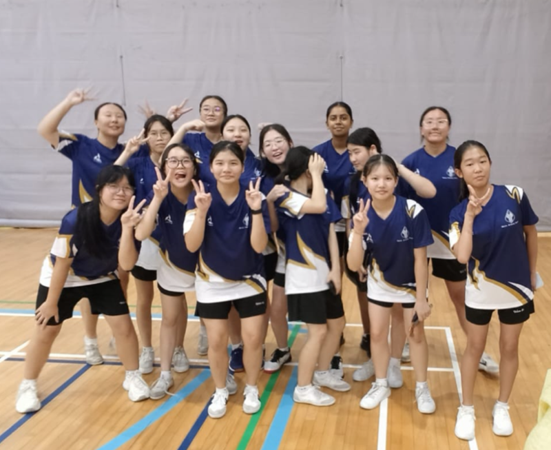 A group of students in badminton uniform pose together on an indoor court, many making peace signs.