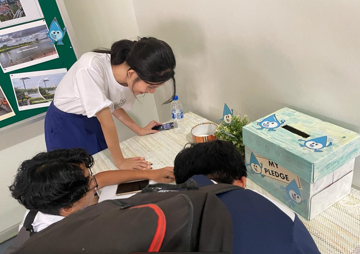 A girl leans over a table showing her phone to two seated students, with a decorated "My Pledge" box nearby.