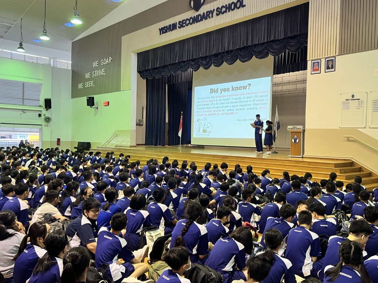 Students in blue uniforms sit on a hall floor at Yishun Secondary School facing a stage with a Did You Know slideshow.