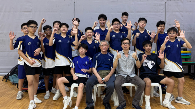 Group of boys students in Badminton uniforms posing with coach and cca teacher, waving and giving thumbs up indoors.