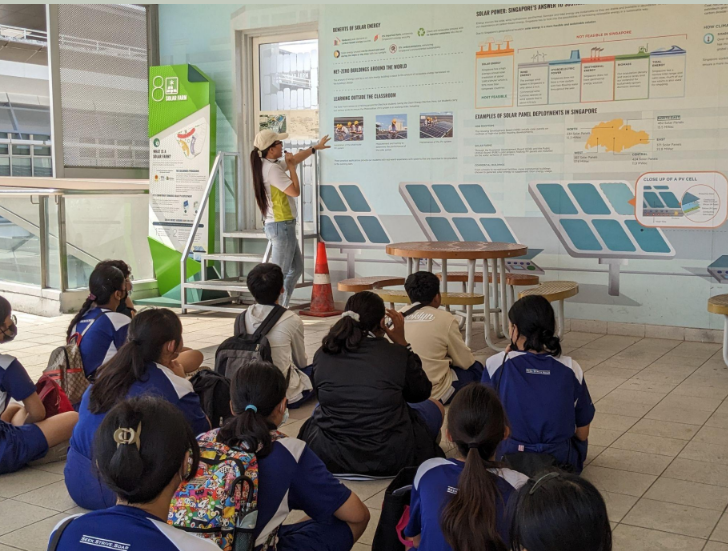 A guide presents a solar power infographic mural to students in blue uniforms seated on the floor.
