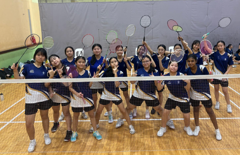 A group of girls in badminton uniforms hold up badminton rackets and pose at a net on an indoor court.