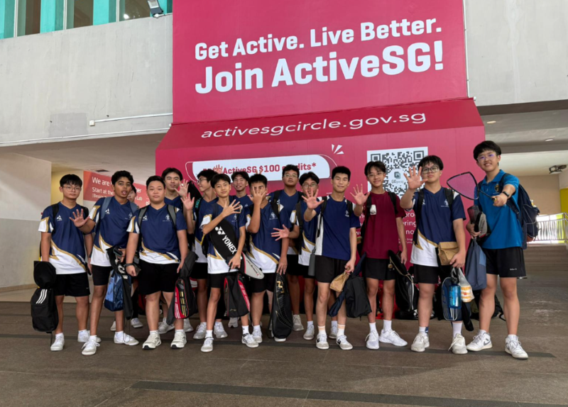 A group of teenage boys in sports uniforms wave and pose in front of a pink ActiveSG banner.