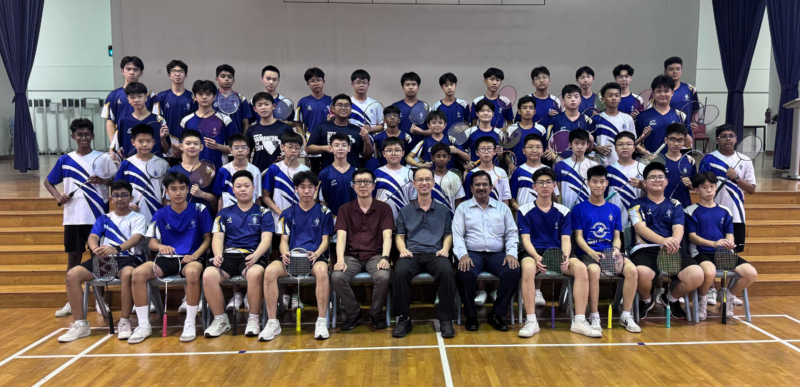 A large group of youth badminton players in blue uniforms pose with three adult coaches on an indoor court.