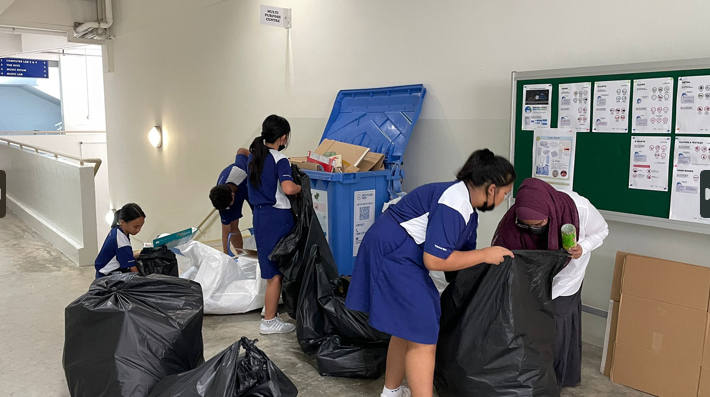 Students in blue uniforms sorting trash bags and recyclables near a large blue recycling bin indoors.