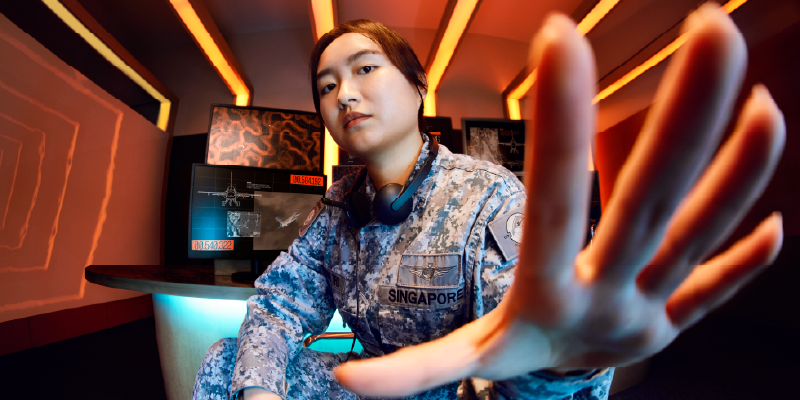 AIRIX in uniform extends her hand extended toward the viewer in a room with screens.