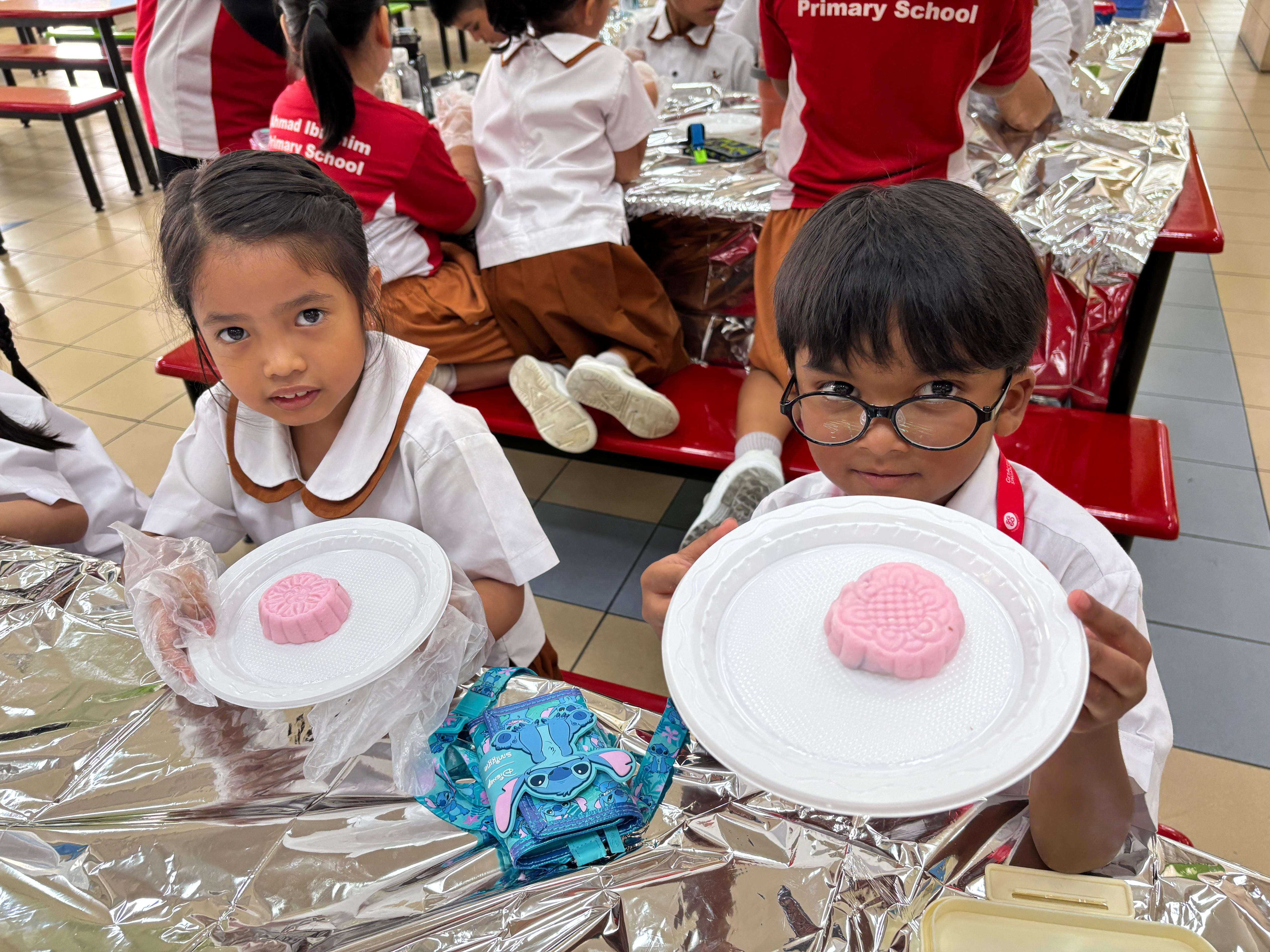 Two young primary school students in uniforms hold white plates each displaying a pink mooncake.