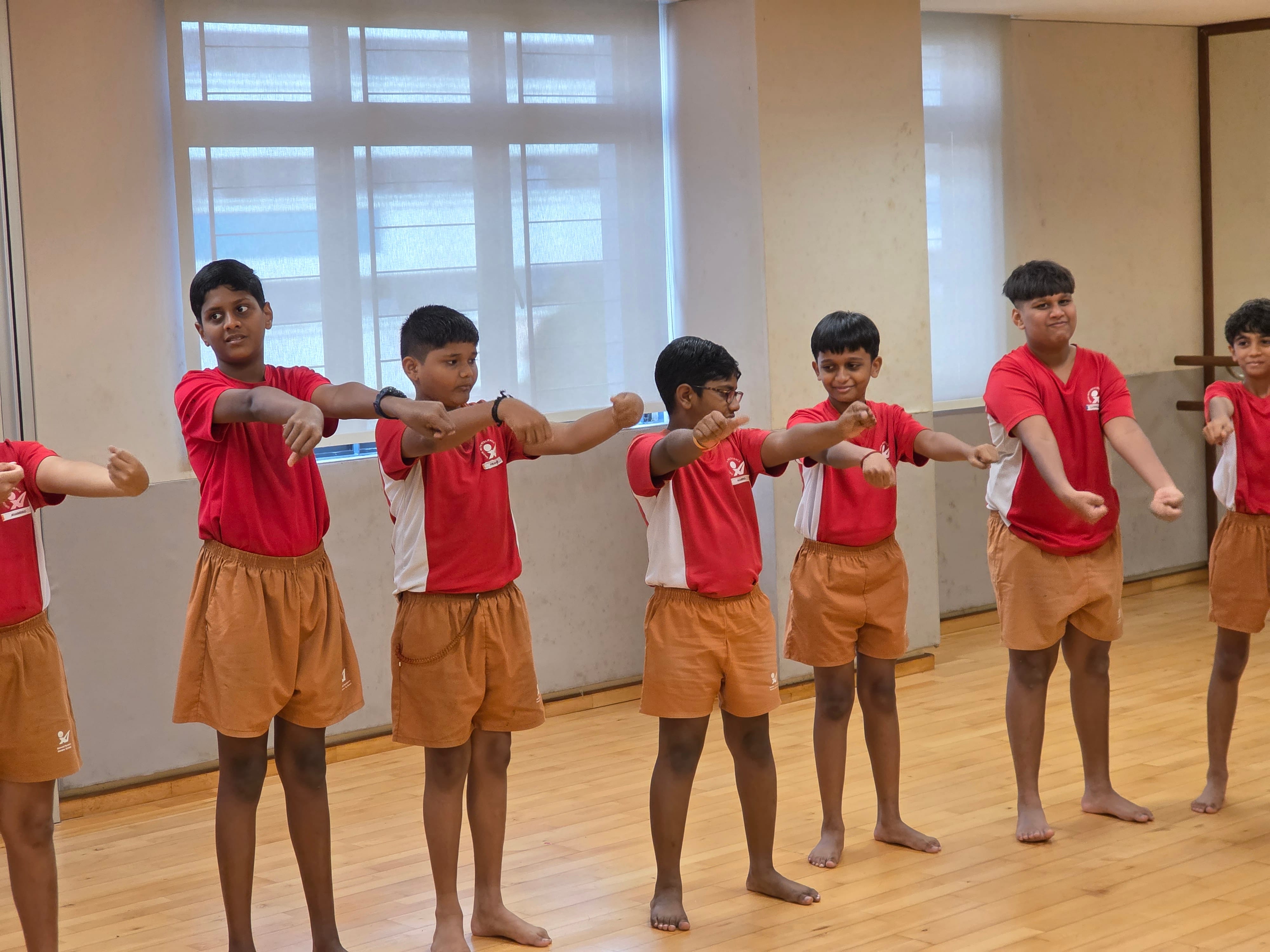 Six boys in red shirts and tan shorts stand barefoot in a studio, arms extended forward in a synchronized pose.