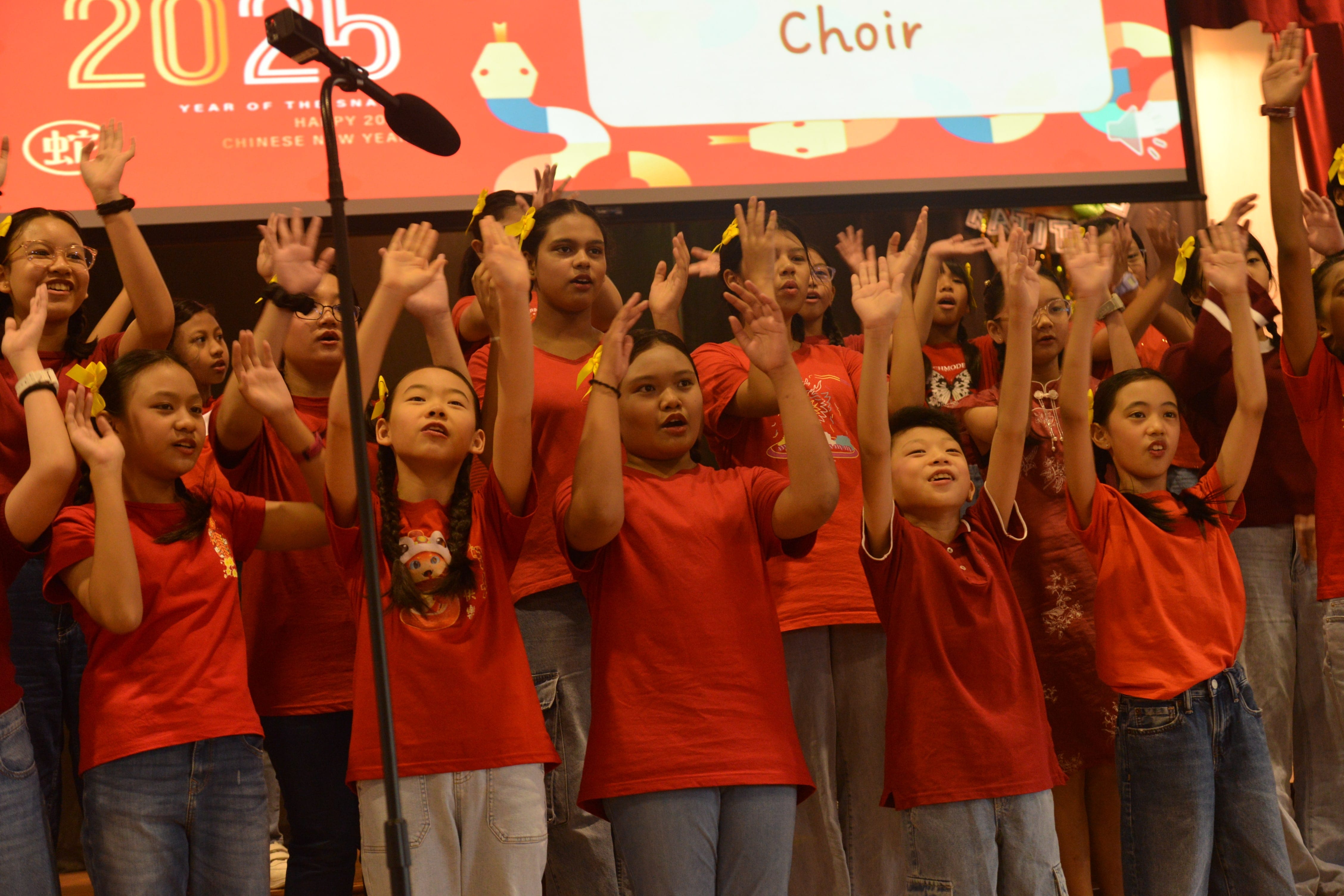 Children in red shirts perform as a choir on stage at a 2025 Chinese New Year celebration, raising their hands.