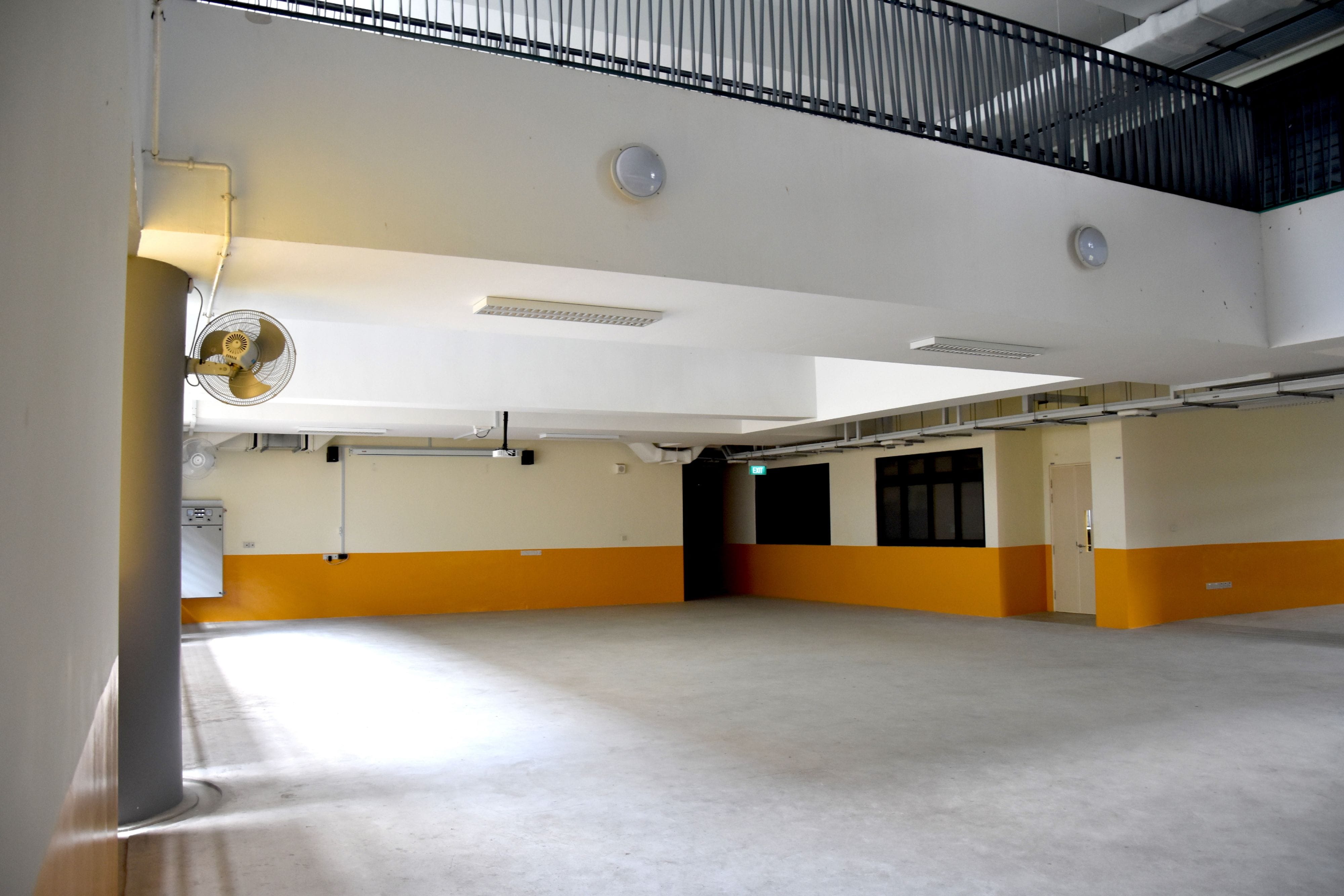 Empty indoor hall with white walls, orange lower stripe, a wall-mounted fan, ceiling lights, and a metal railing above.