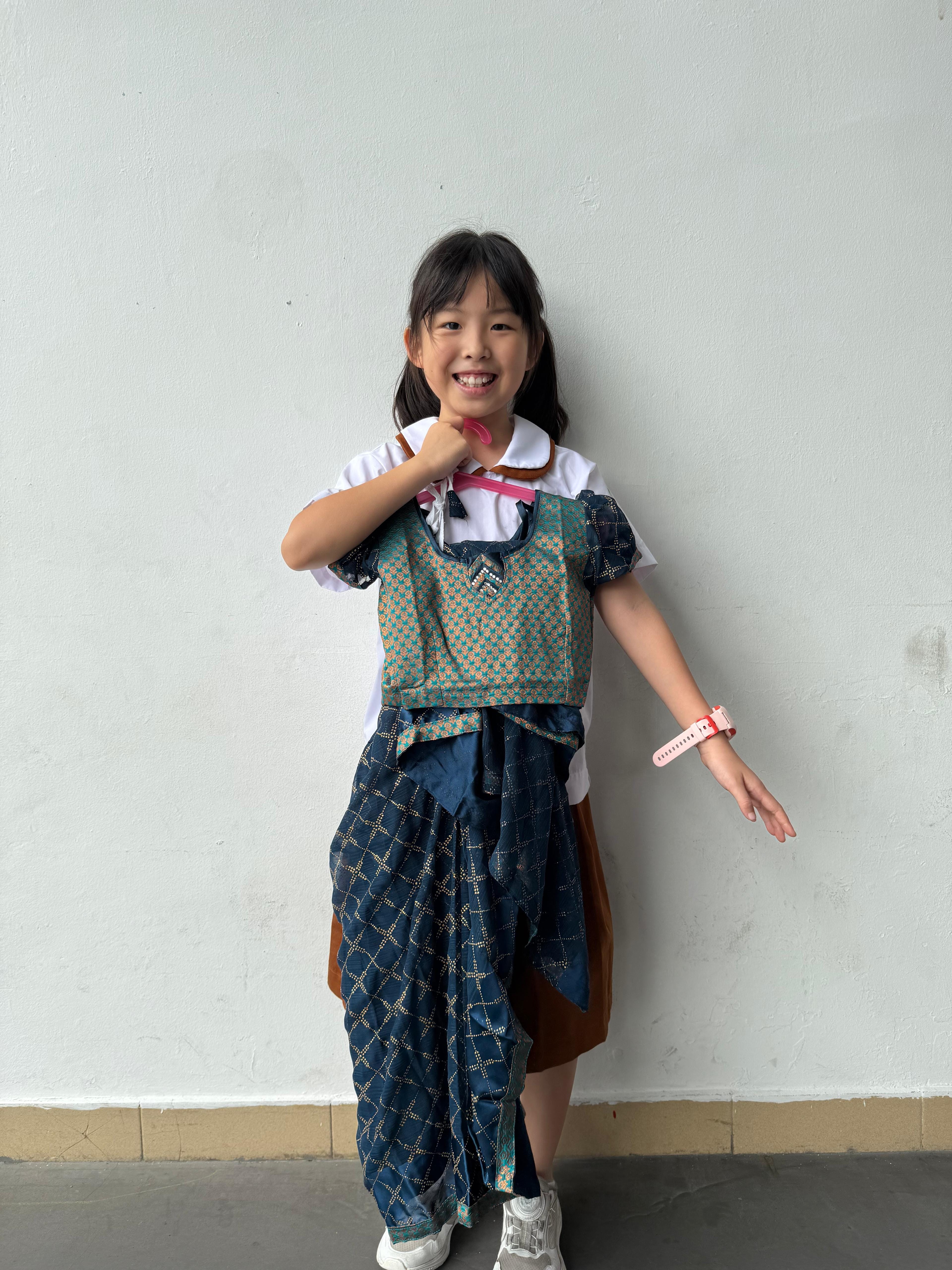 A smiling young girl wears a traditional costume over a white shirt, posing against a light gray wall.