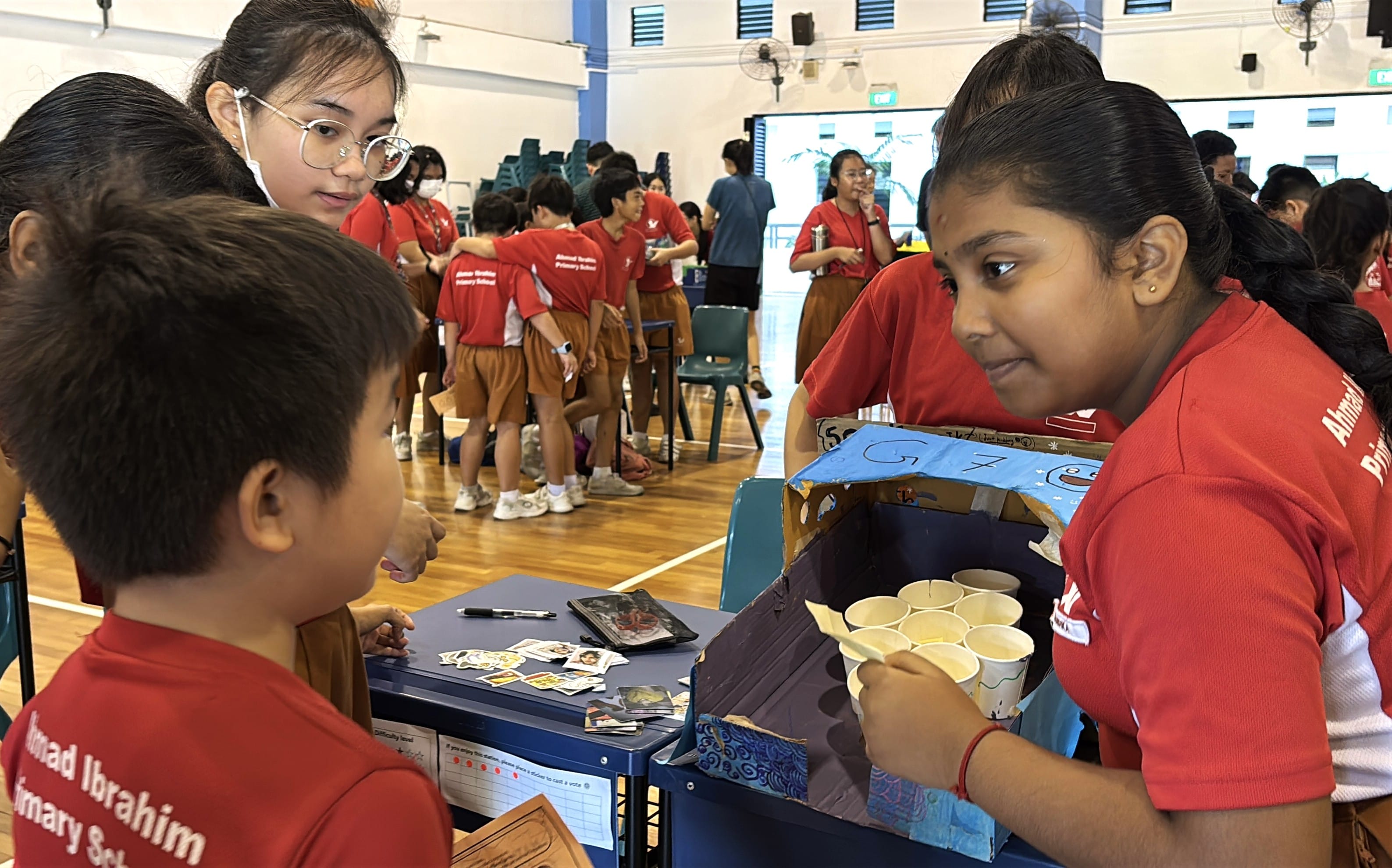 Students in red Ahmad Ibrahim Primary School uniforms at an indoor activity, one holding a box with paper cups.