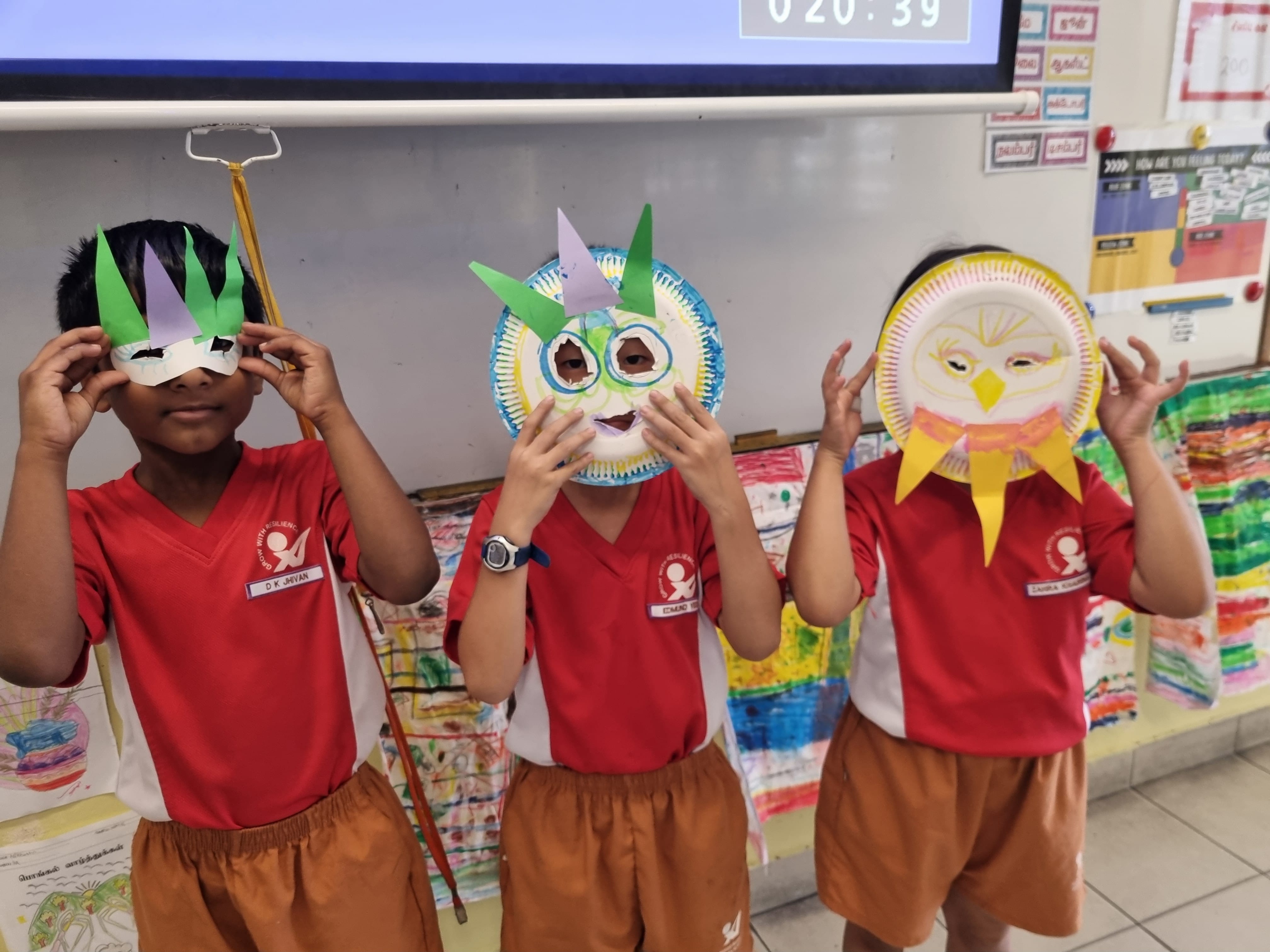 Three children in red school uniforms hold up handmade paper plate owl masks in a colorful classroom.