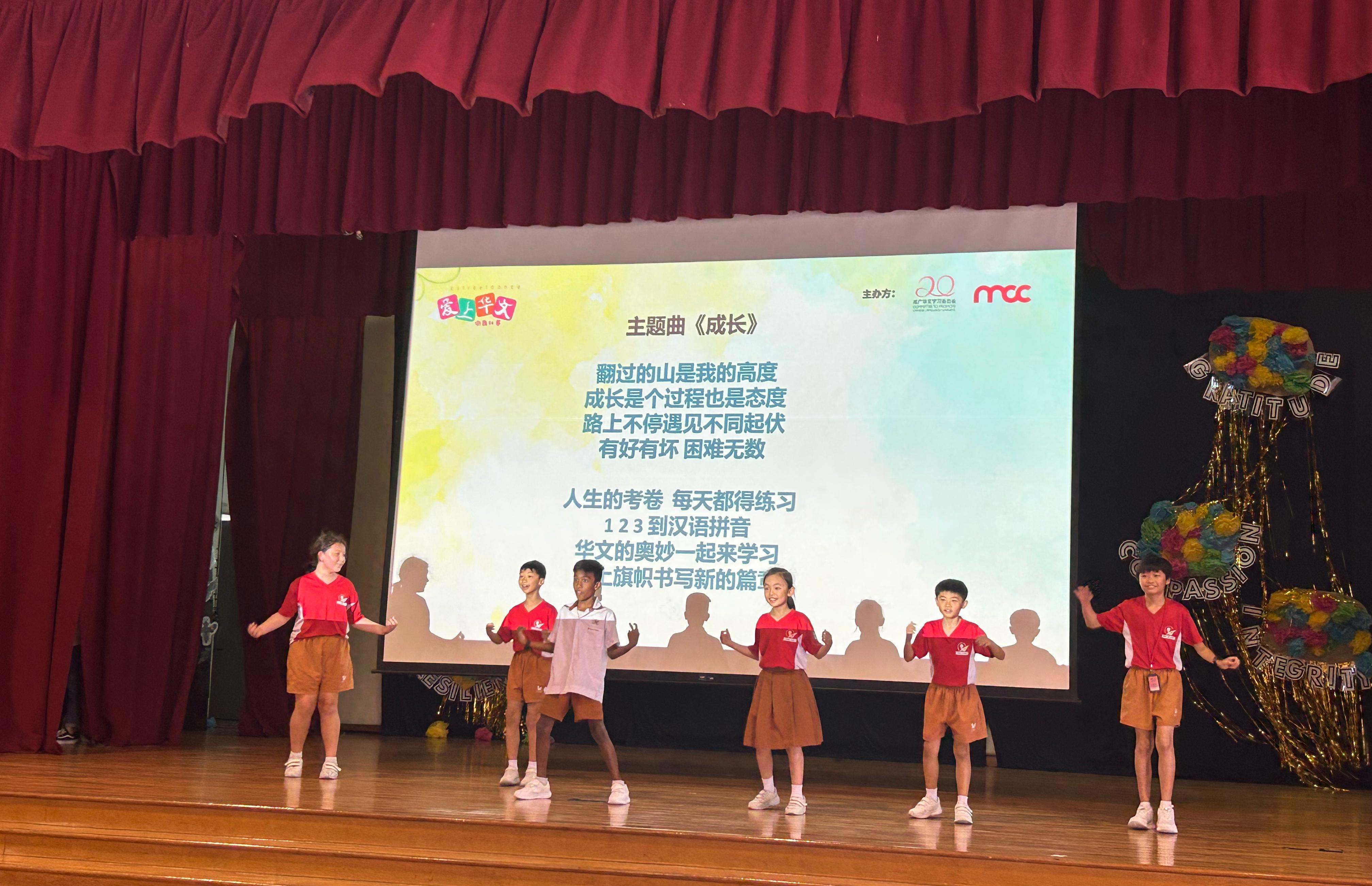 Six children in red uniforms perform on a stage in front of a screen displaying Chinese song lyrics.