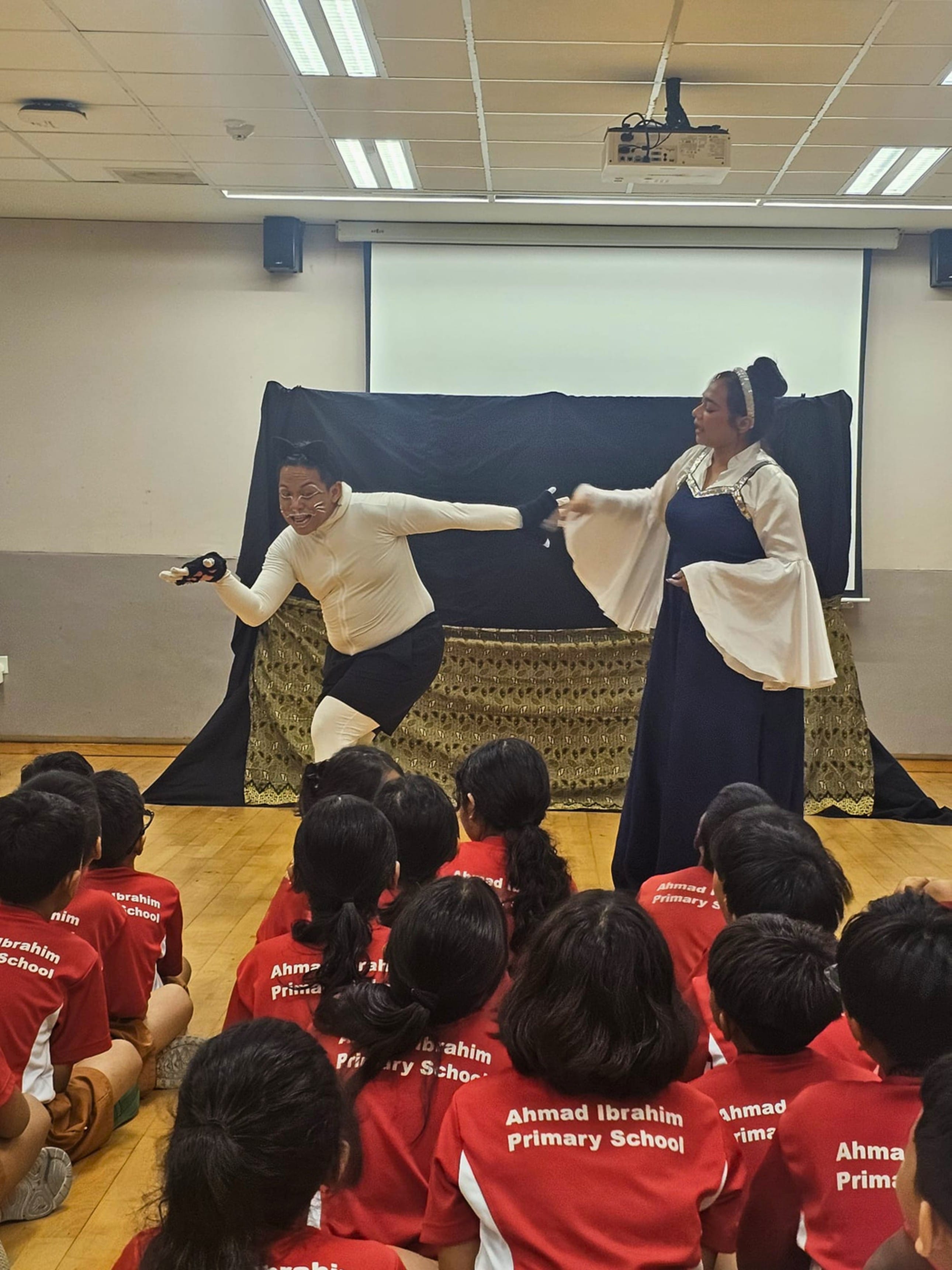 Two costumed performers act on stage before seated children in red Ahmad Ibrahim Primary School uniforms.