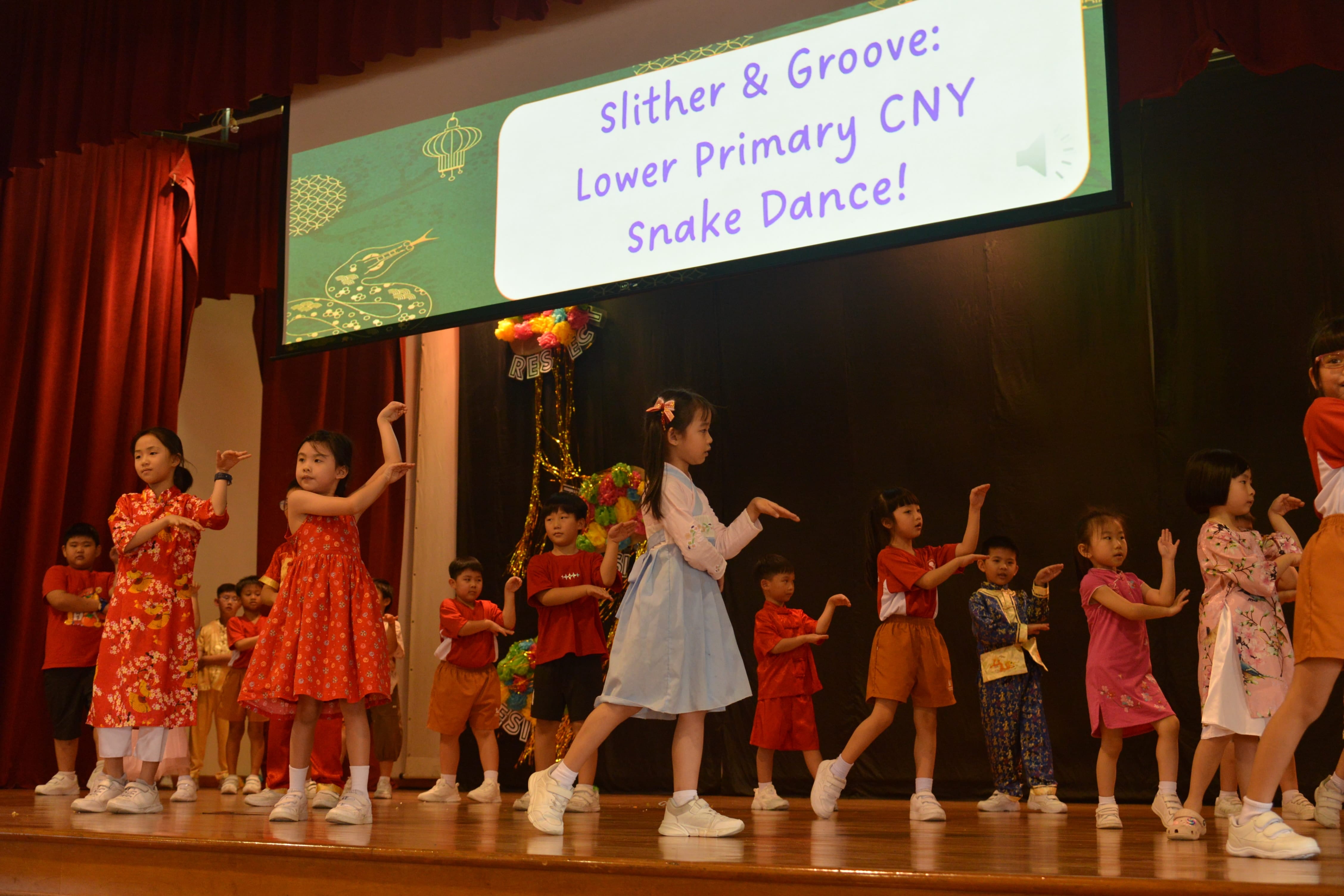 Young children in colorful outfits perform a snake dance on stage at a Lower Primary Chinese New Year event.