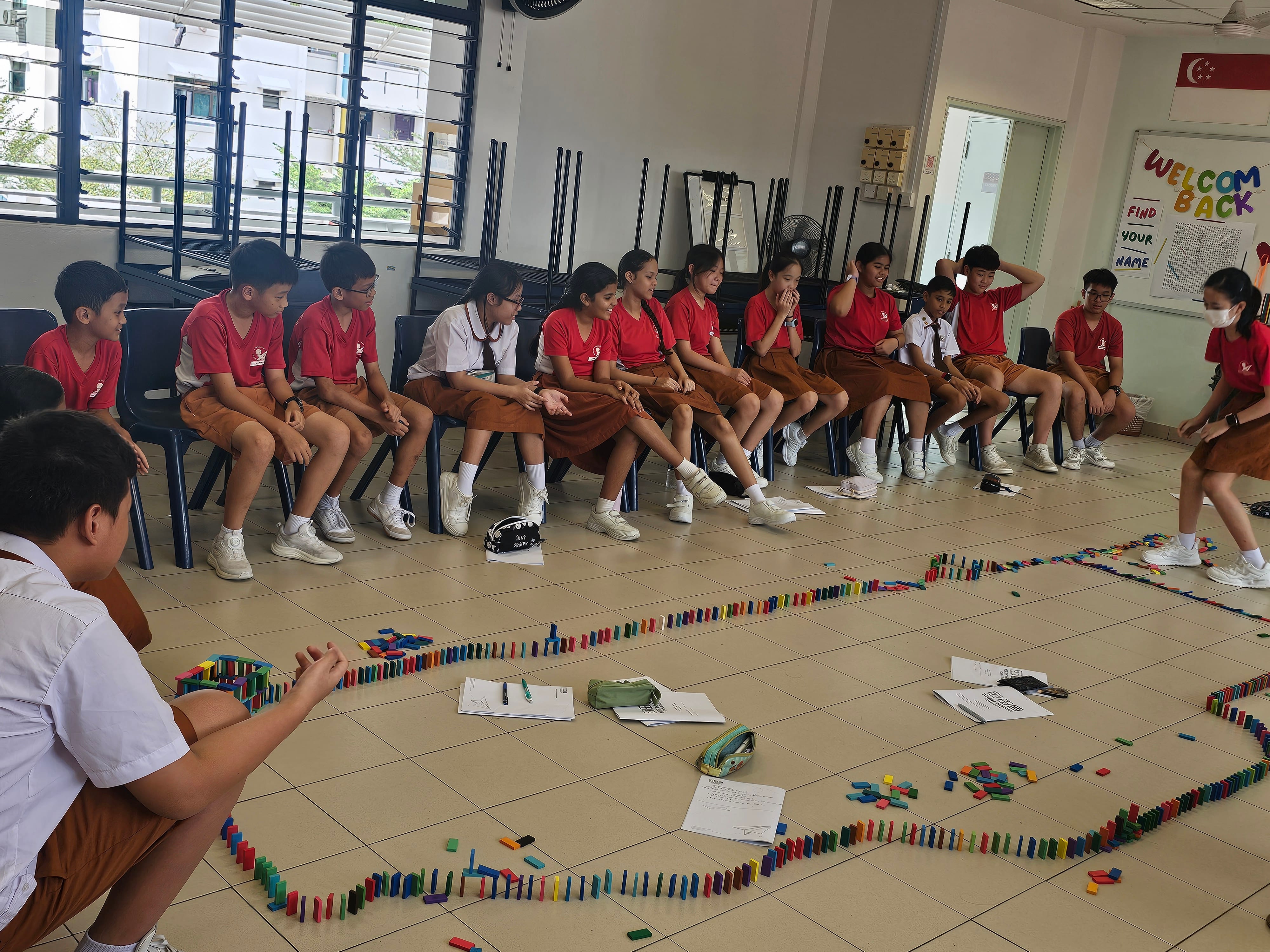 Students in red uniforms sit watching a large colorful domino trail arranged on a classroom floor.