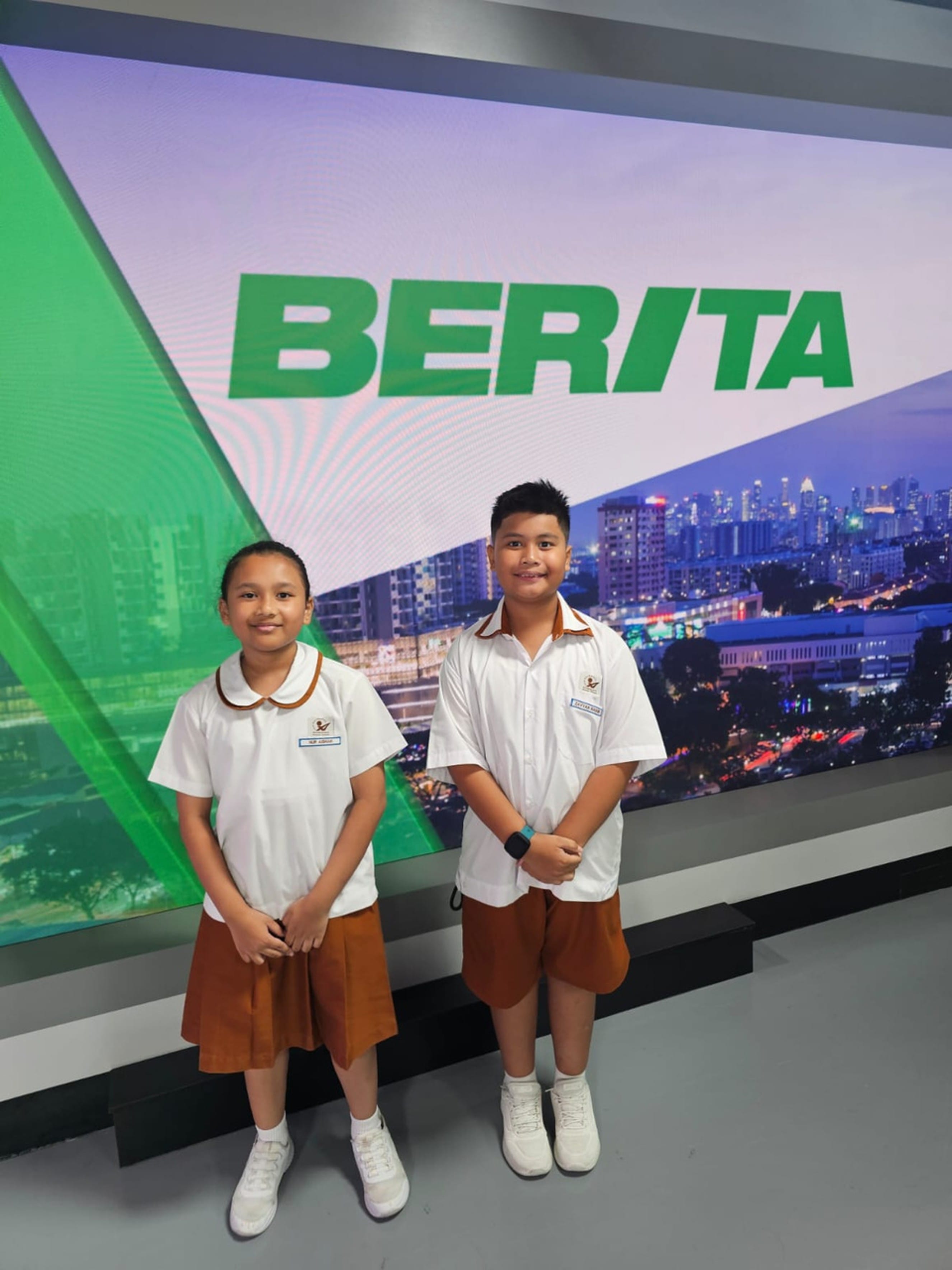 Two children in white and brown school uniforms stand smiling in front of a large screen displaying the word BERITA.