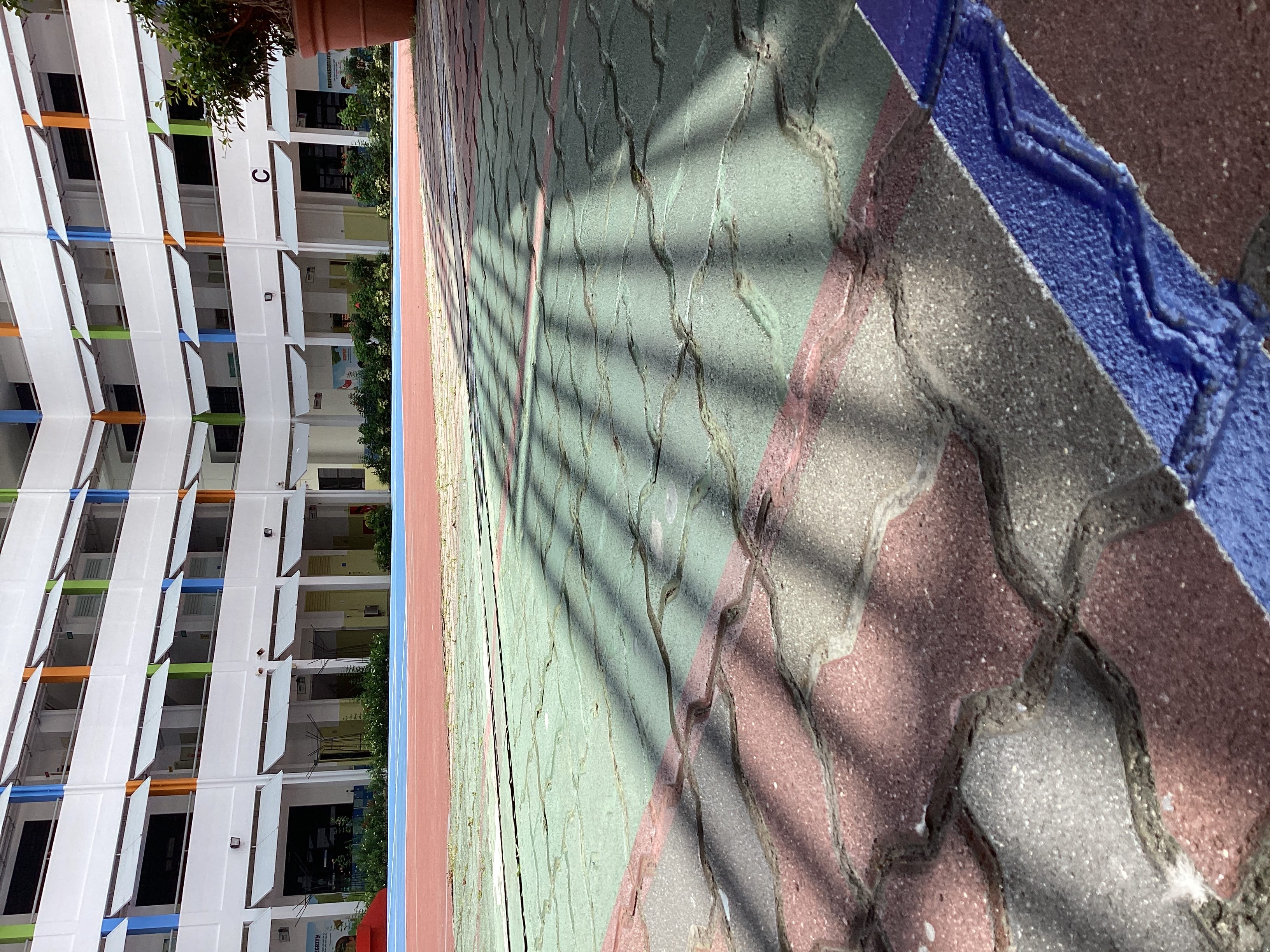 Colorful paved courtyard with red and green sections, fence shadows, and a multi-story white school building behind.