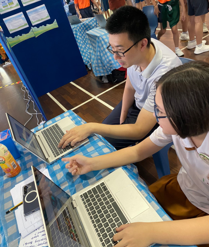 Two students wearing glasses work on laptops at a table covered with a blue checkered cloth at a school fair.