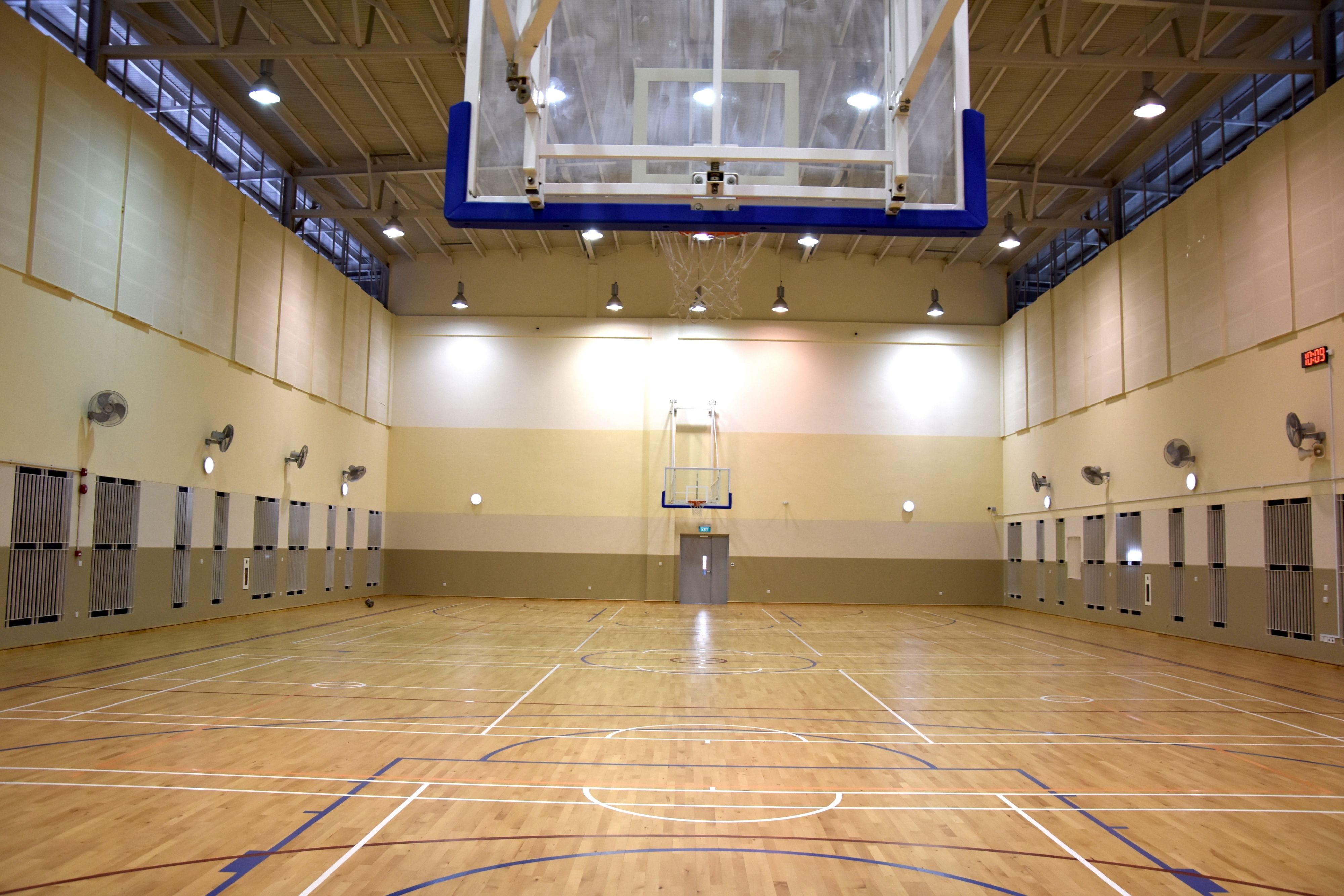 Empty indoor basketball court with wood floor, painted lines, two hoops, wall fans, and overhead lights.