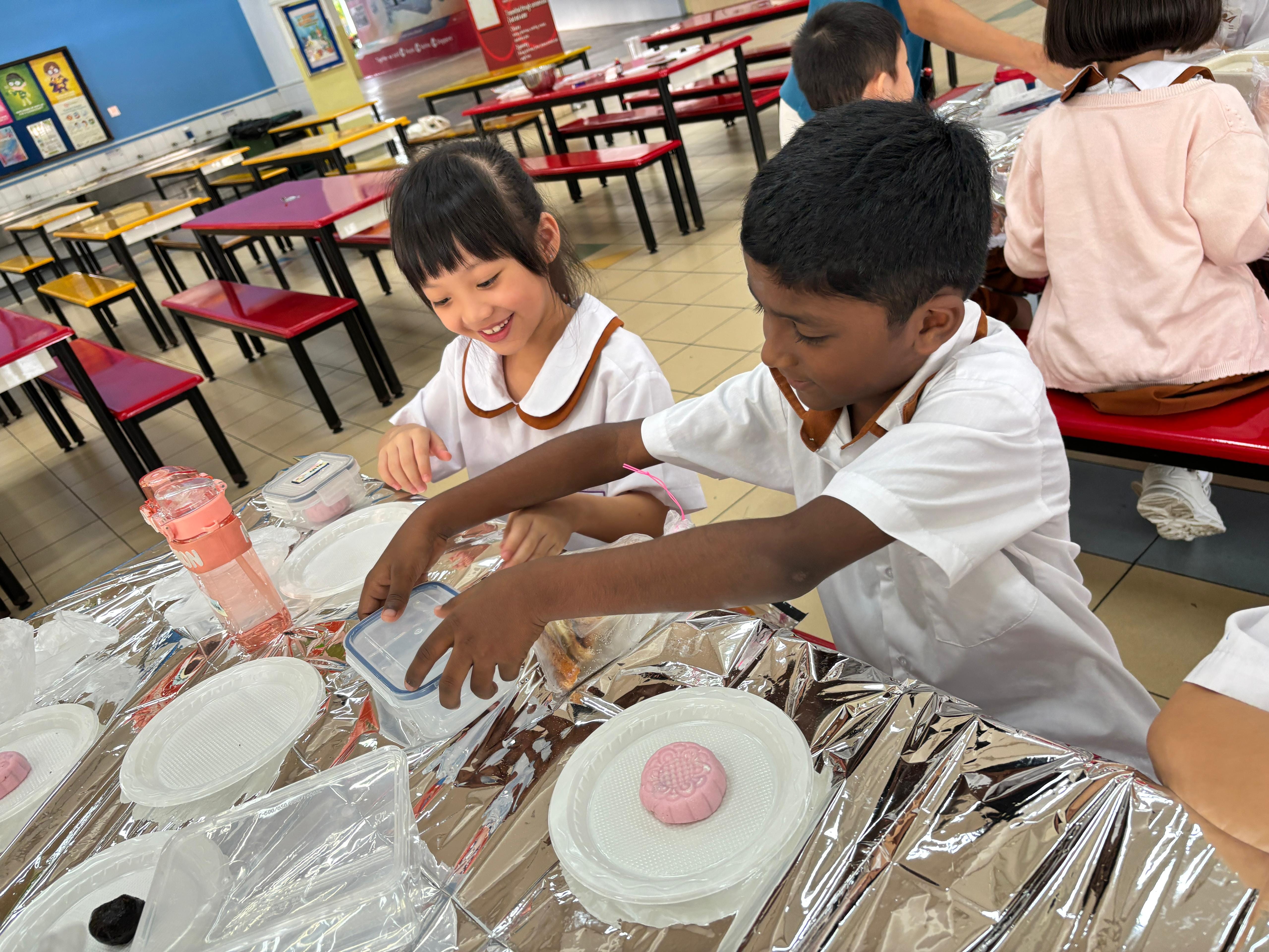 Two school children in white uniforms make pink mooncakes at a foil-covered table in a school canteen.