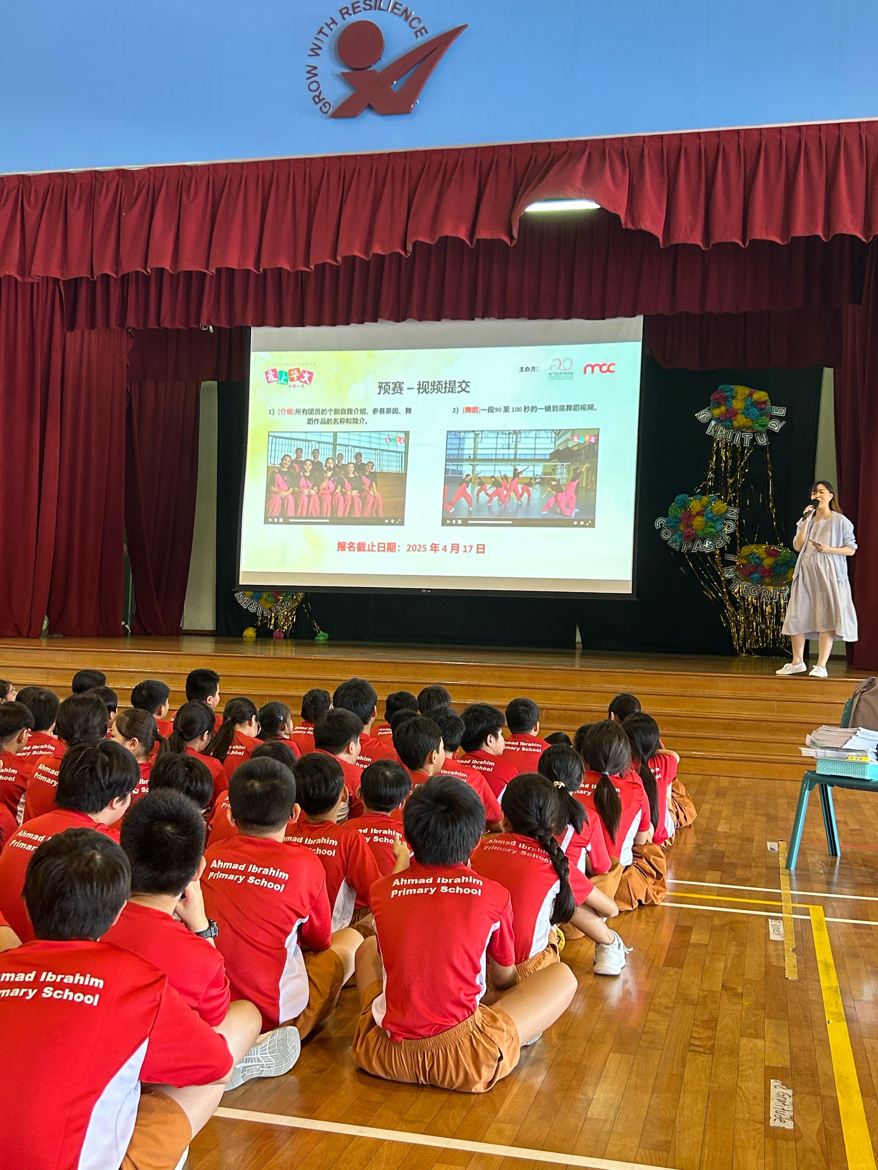 Students in red Ahmad Ibrahim Primary School shirts sit on a gym floor watching a Chinese-language slideshow presentation on stage.